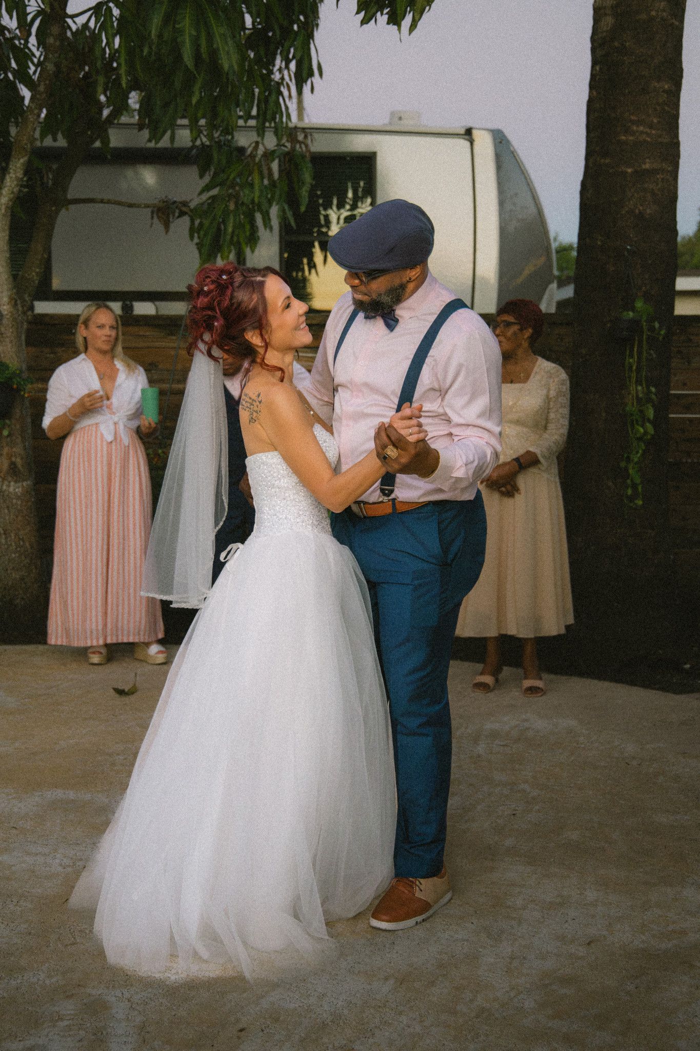 A couple in wedding attire shares a romantic dance moment outdoors under palm trees at a reception venue.