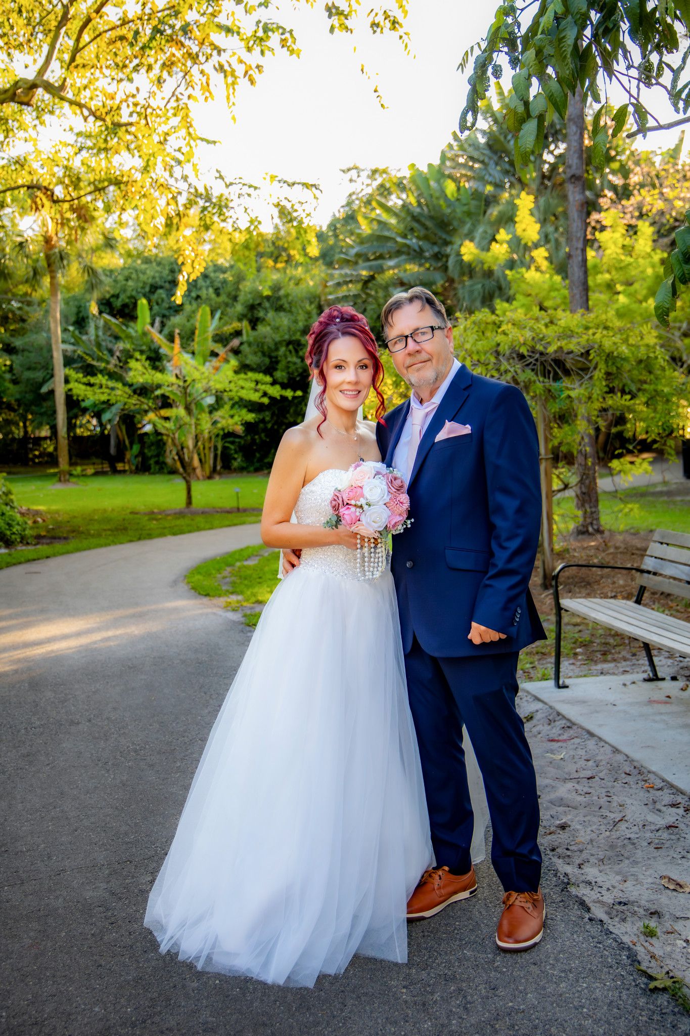 A couple poses together on a paved path in a park during golden hour with autumn foliage in the background.