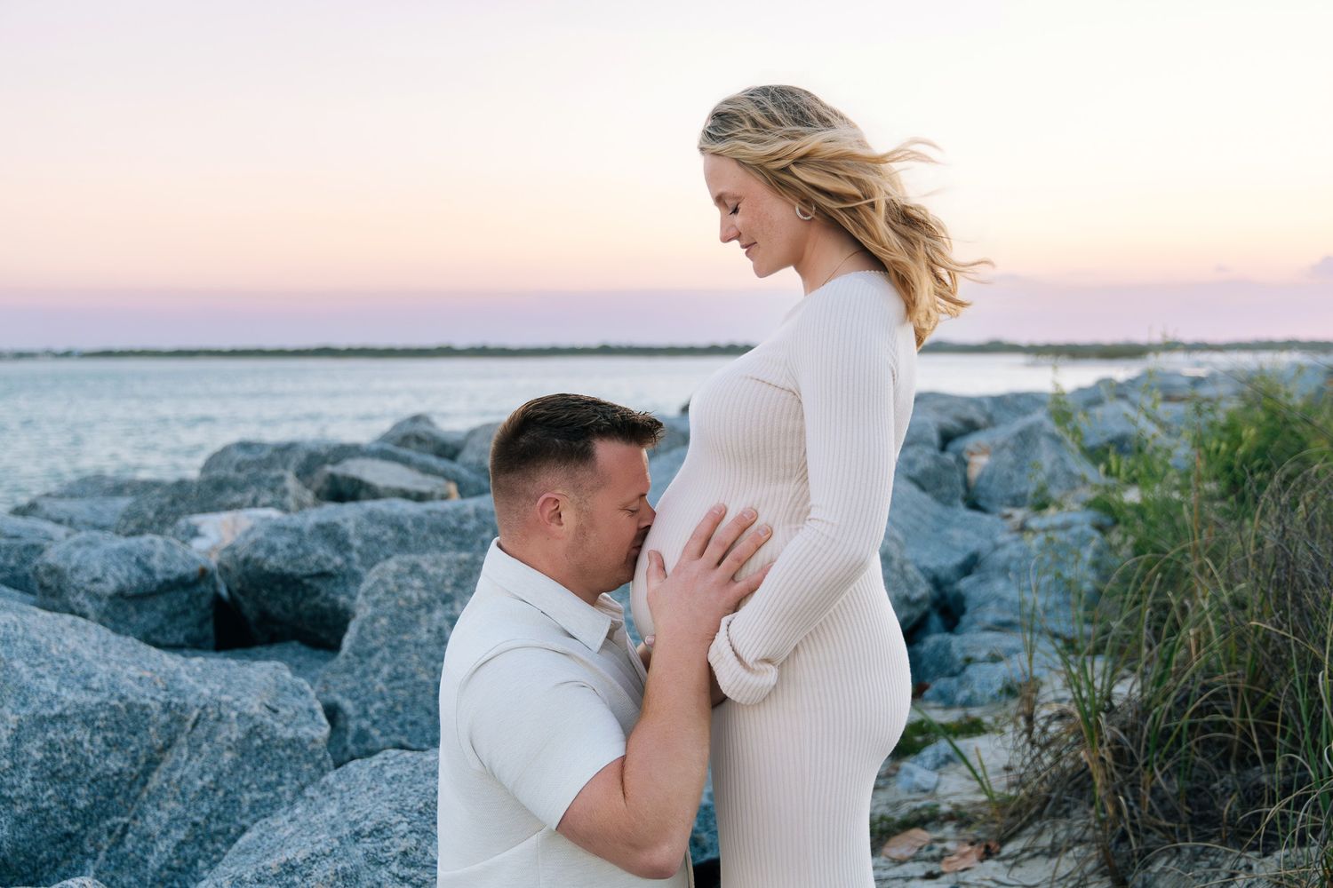 Romantic coastal engagement photo session with couple in white outfits on rocky beach at sunset.
