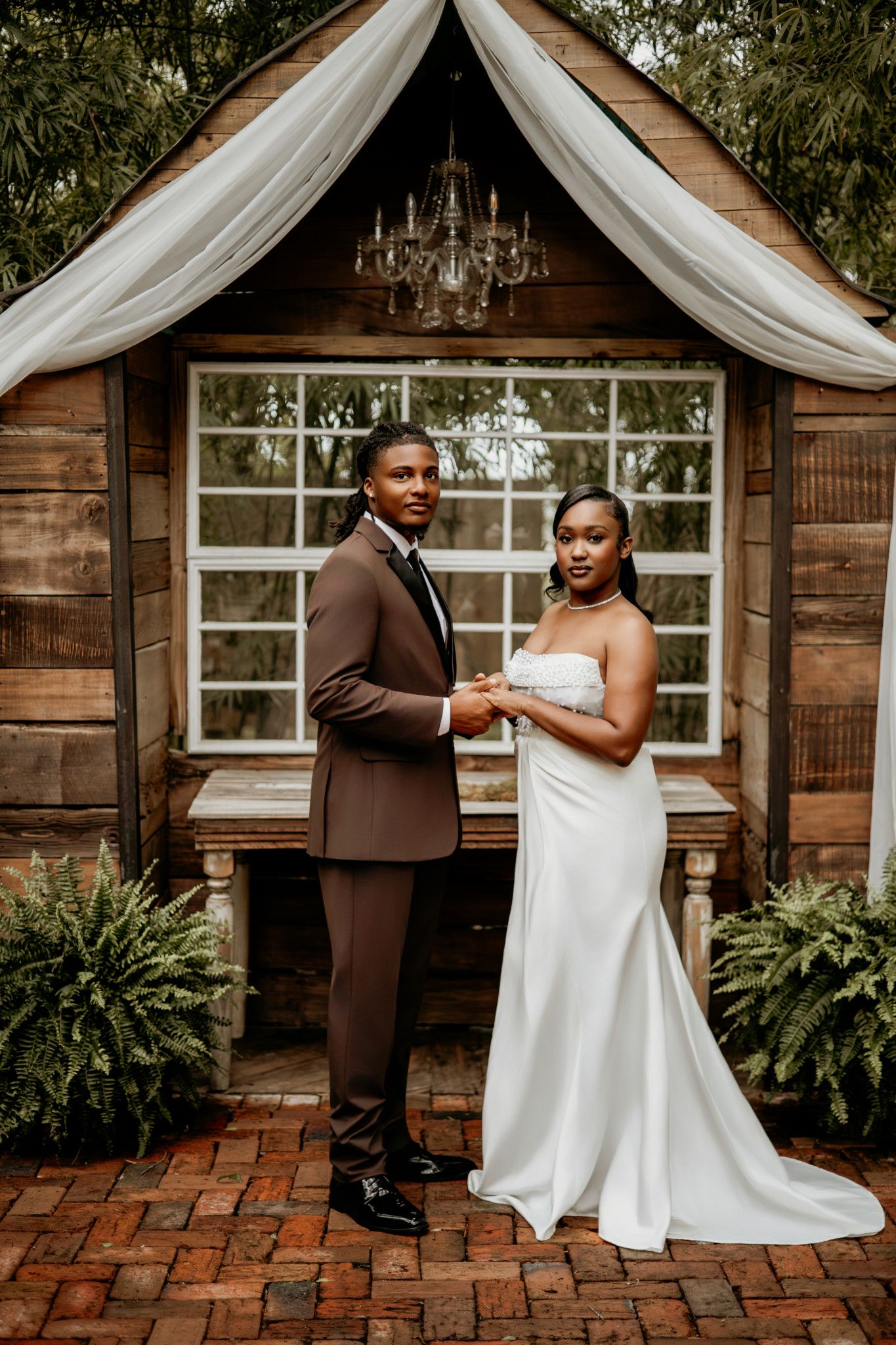 A couple shares an intimate dance moment on a brick patio outside a rustic wooden building with white drapes.