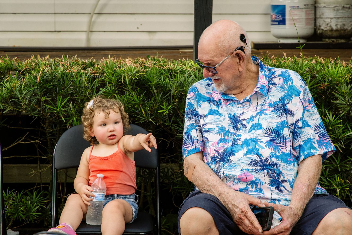 Elderly man in floral shirt sits with a toddler girl in orange tank top on black chairs outdoors.