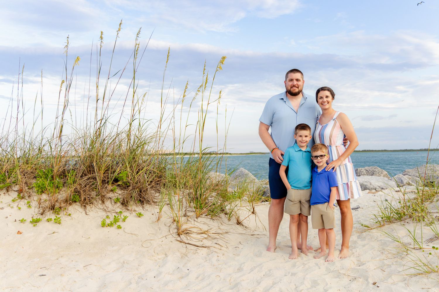 A family of four poses on a sandy beach with sea oats and calm water in the background.