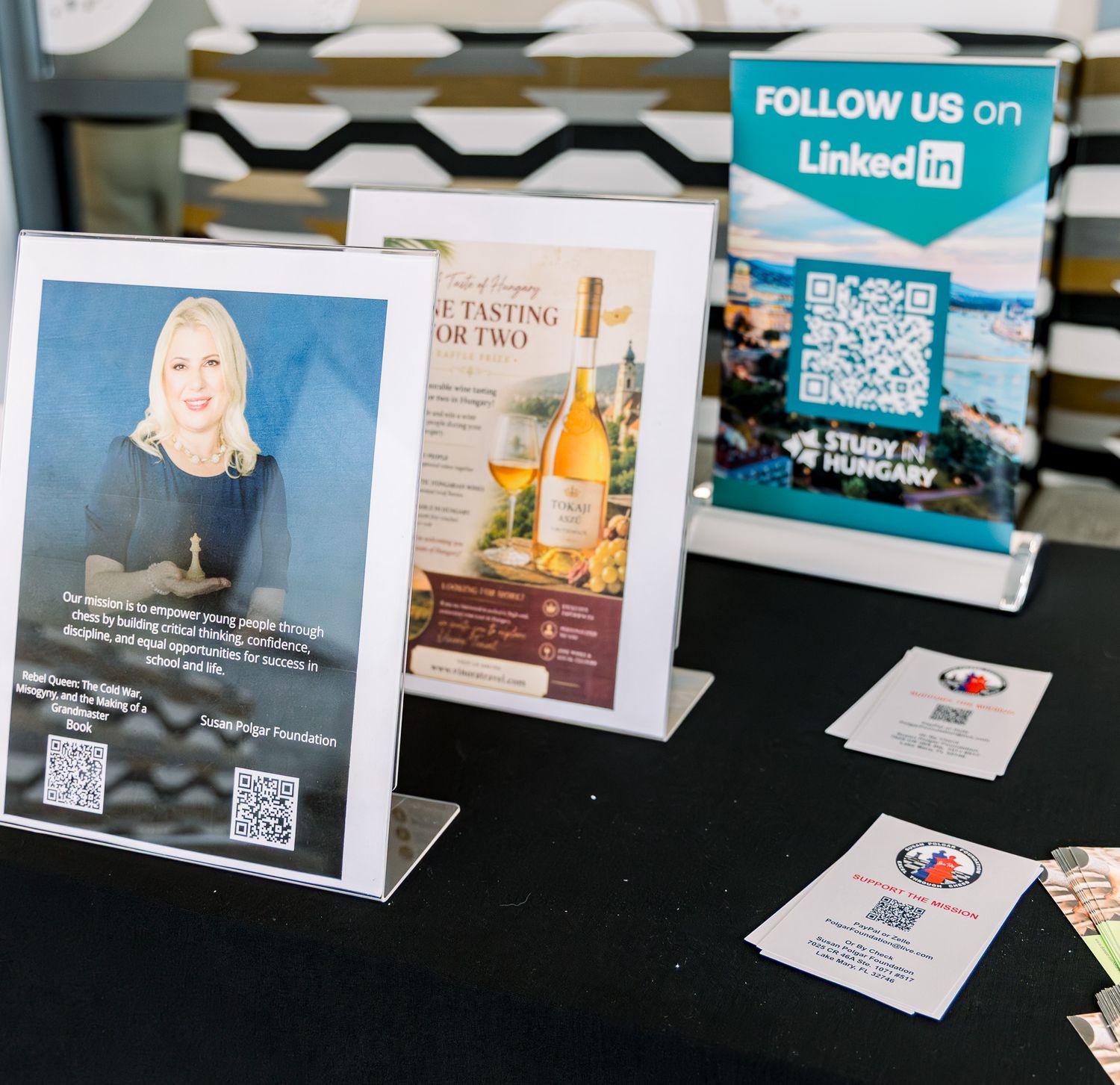 A trade show table displays LinkedIn QR code sign, wine tasting brochure, and business cards on black tablecloth.