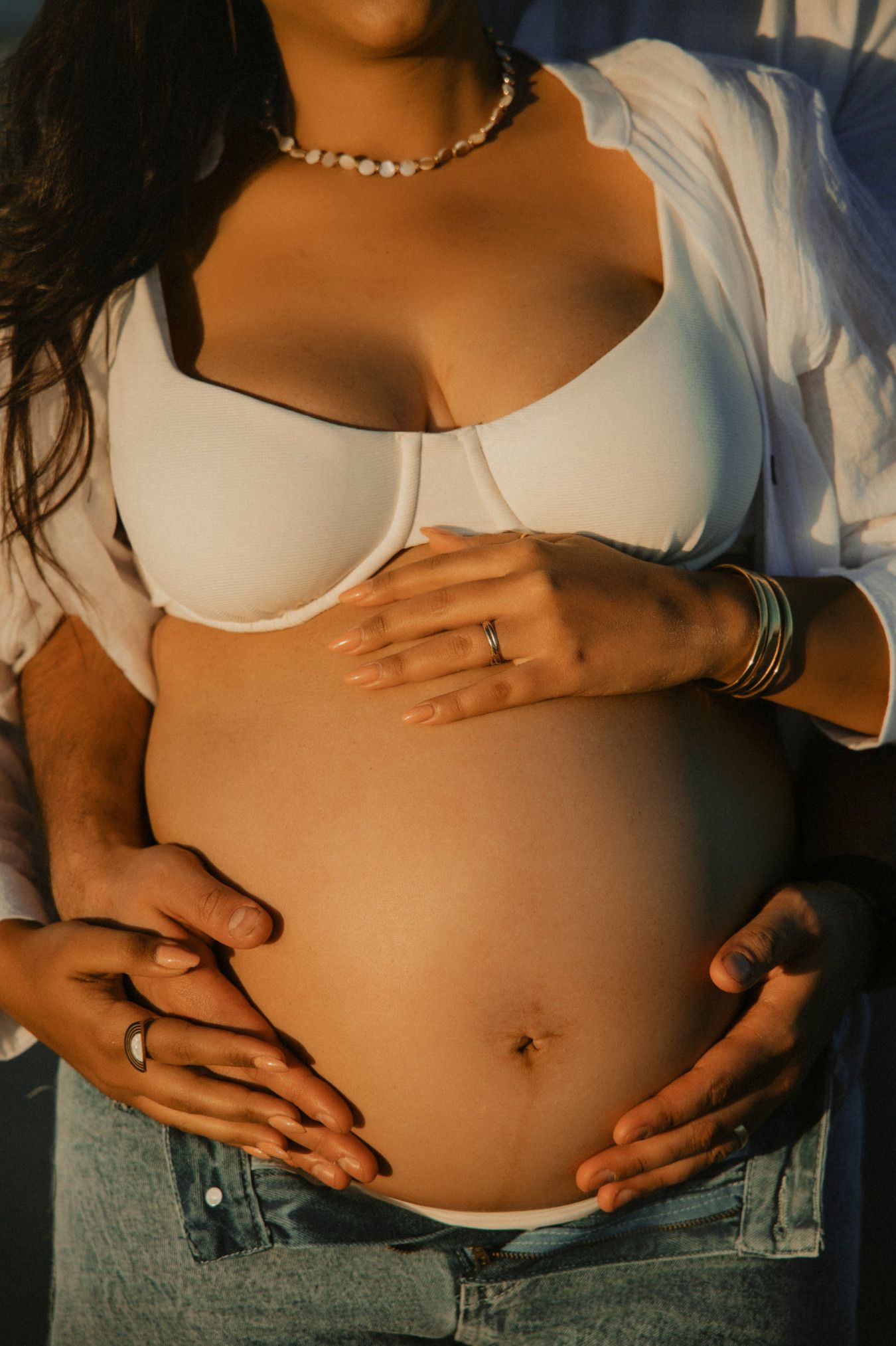 Close-up of pregnant couple cradling baby bump, woman wearing white bra, jeans, pearl necklace, and gold bracelets.