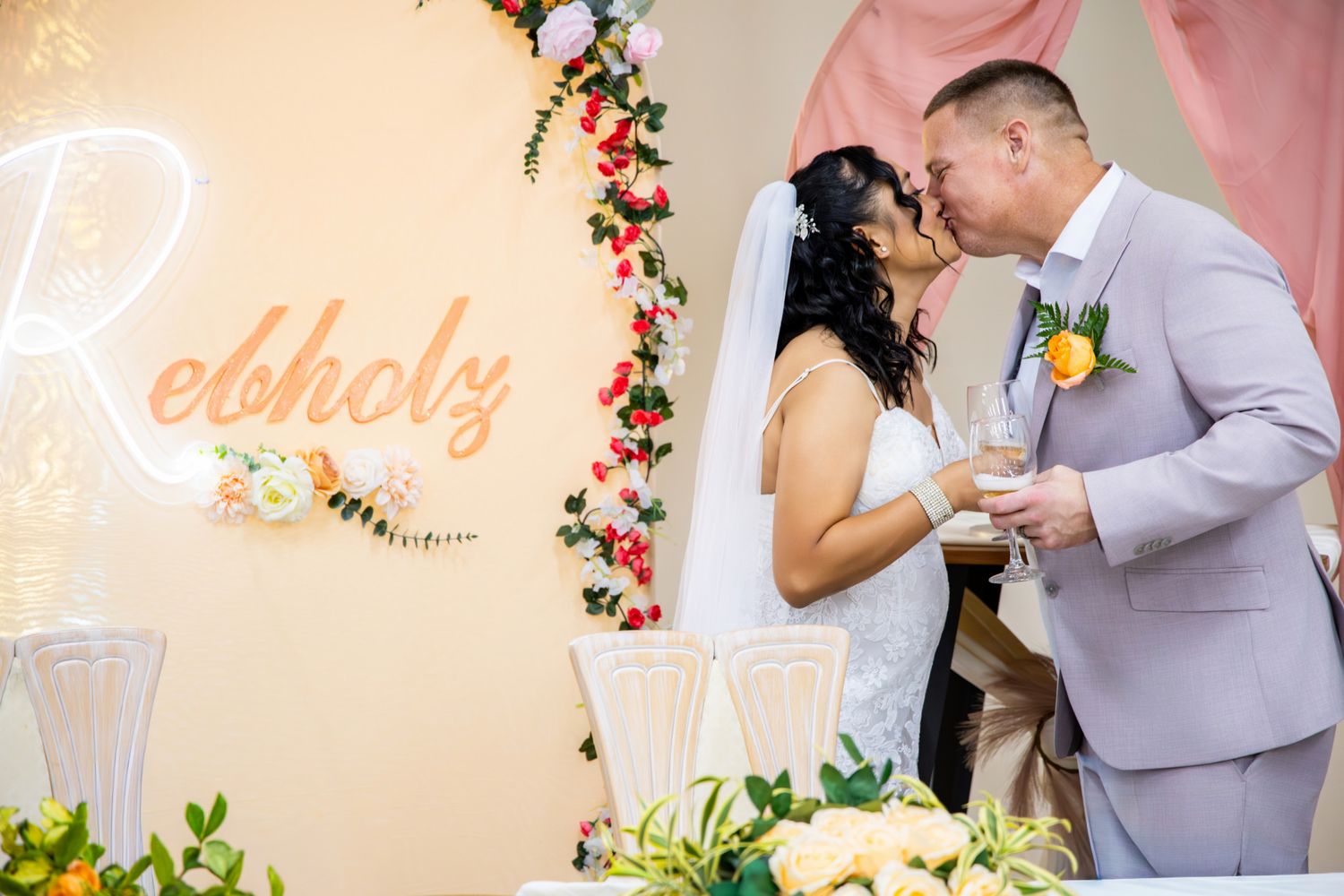 Bride and groom share a kiss at wedding altar decorated with floral arrangements and a glowing neon sign.
