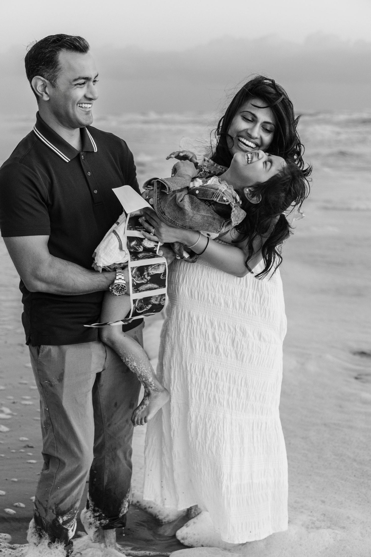 Black and white photo of a smiling couple playfully holding a laughing child at the water's edge on a beach.