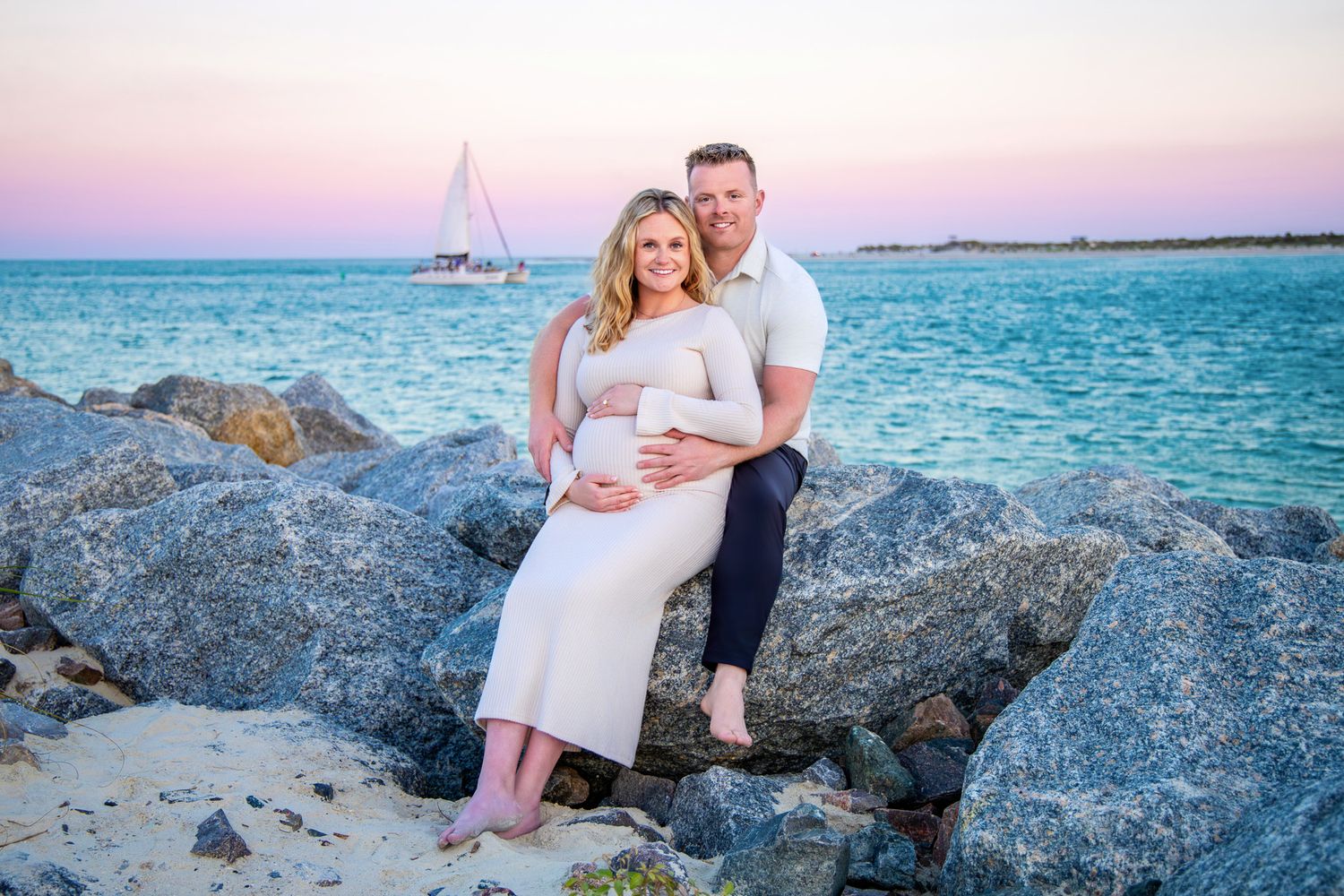 Romantic coastal photoshoot at sunset with couple sitting on rocky beach beside turquoise ocean waters.