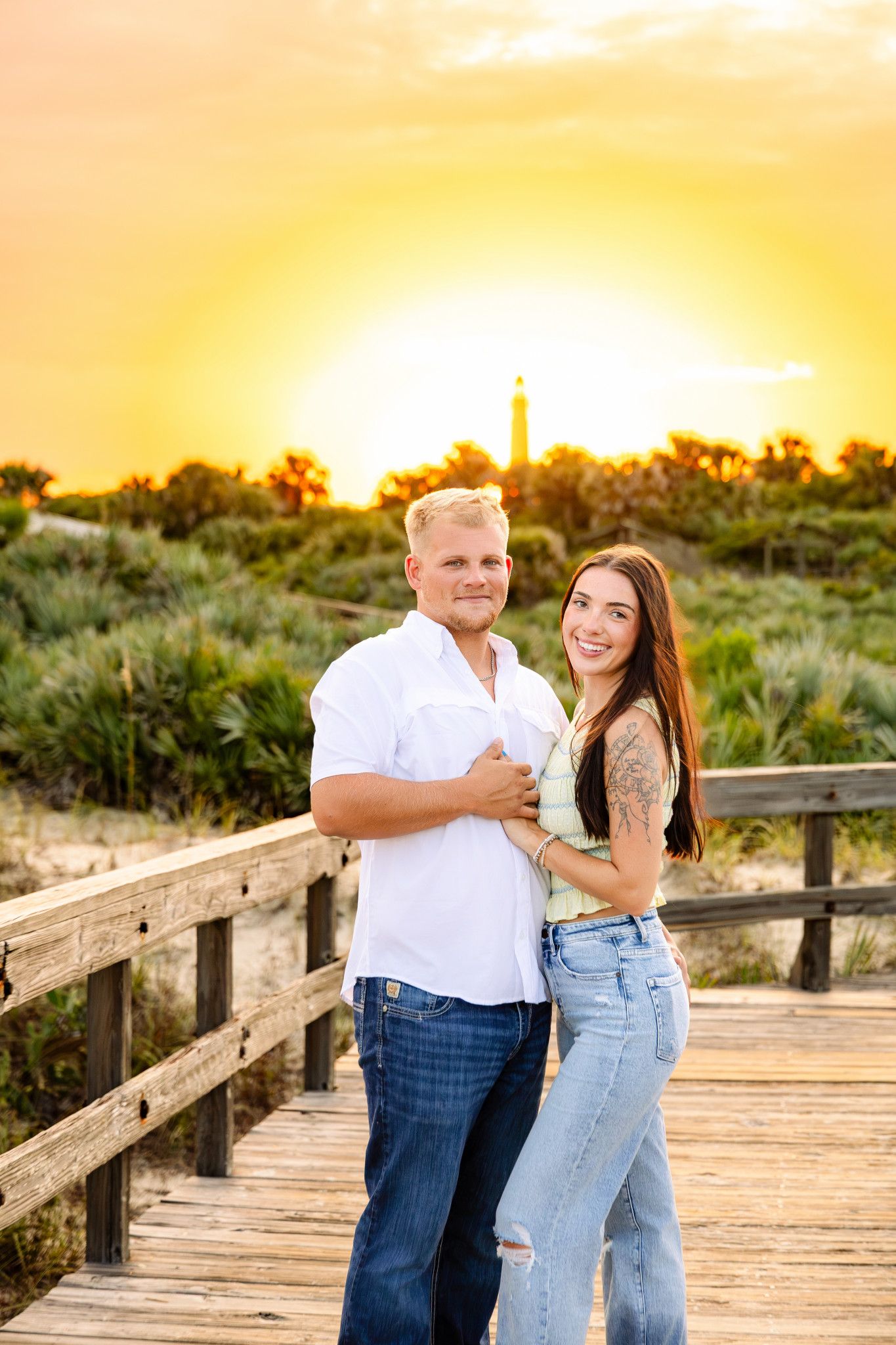 Man and woman posing together on a beach boardwalk during a vibrant golden sunset with a distant lighthouse.
