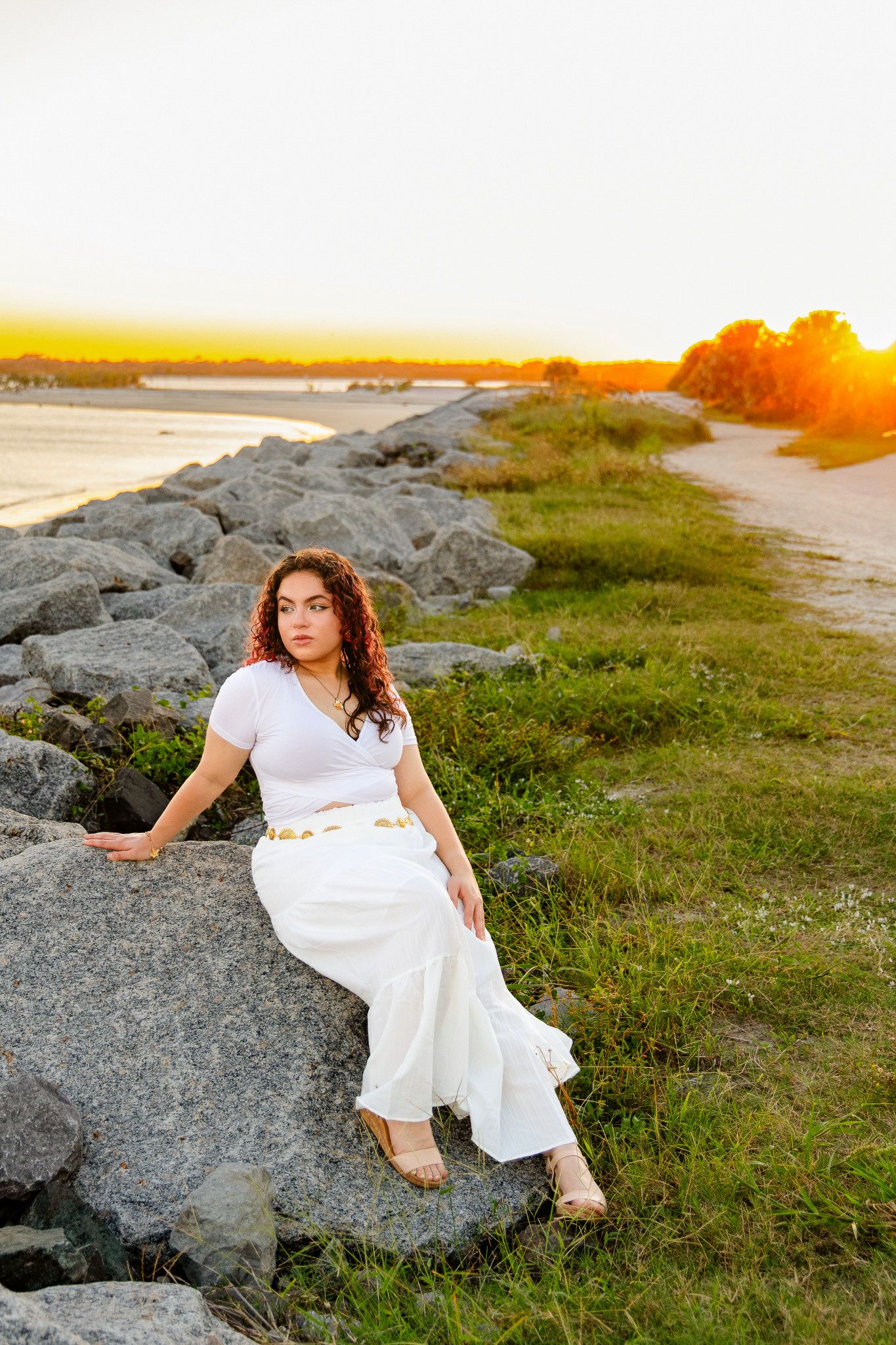 Young woman in a white top and flowing white skirt sits on coastal rocks at sunset, with grassy shoreline and golden light behind her.