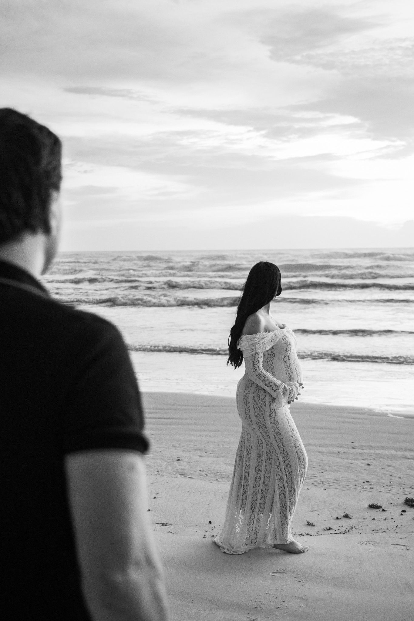 Black and white photo of pregnant woman in lace gown standing on beach at sunset with man in foreground.