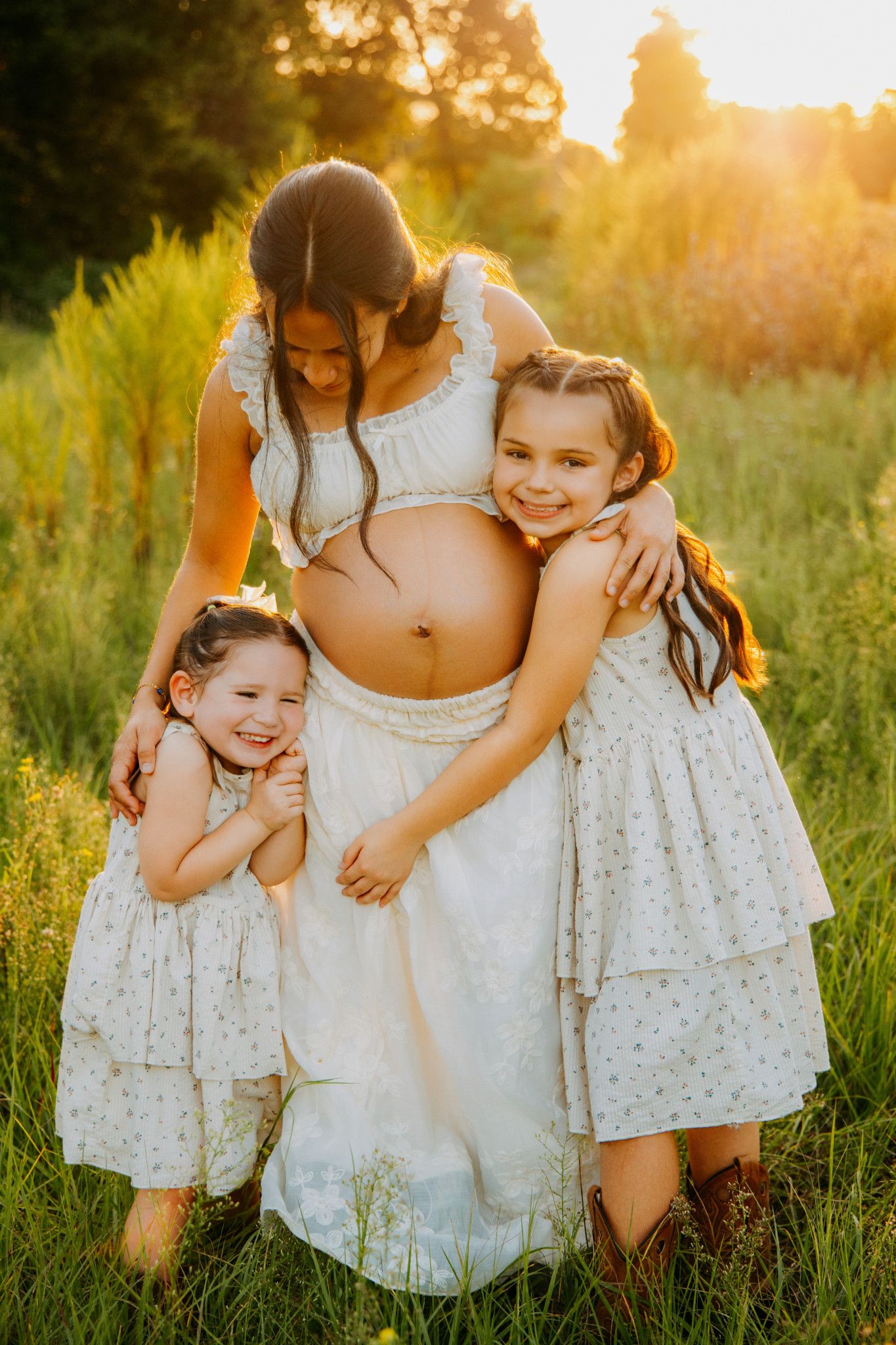 Pregnant mom in white dress embraced by two young daughters in sunny green field at golden hour.