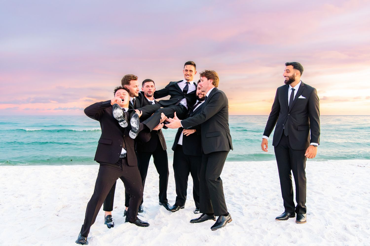 Groomsmen in black suits laughing and celebrating on a sandy beach at sunset with colorful pink and blue sky.