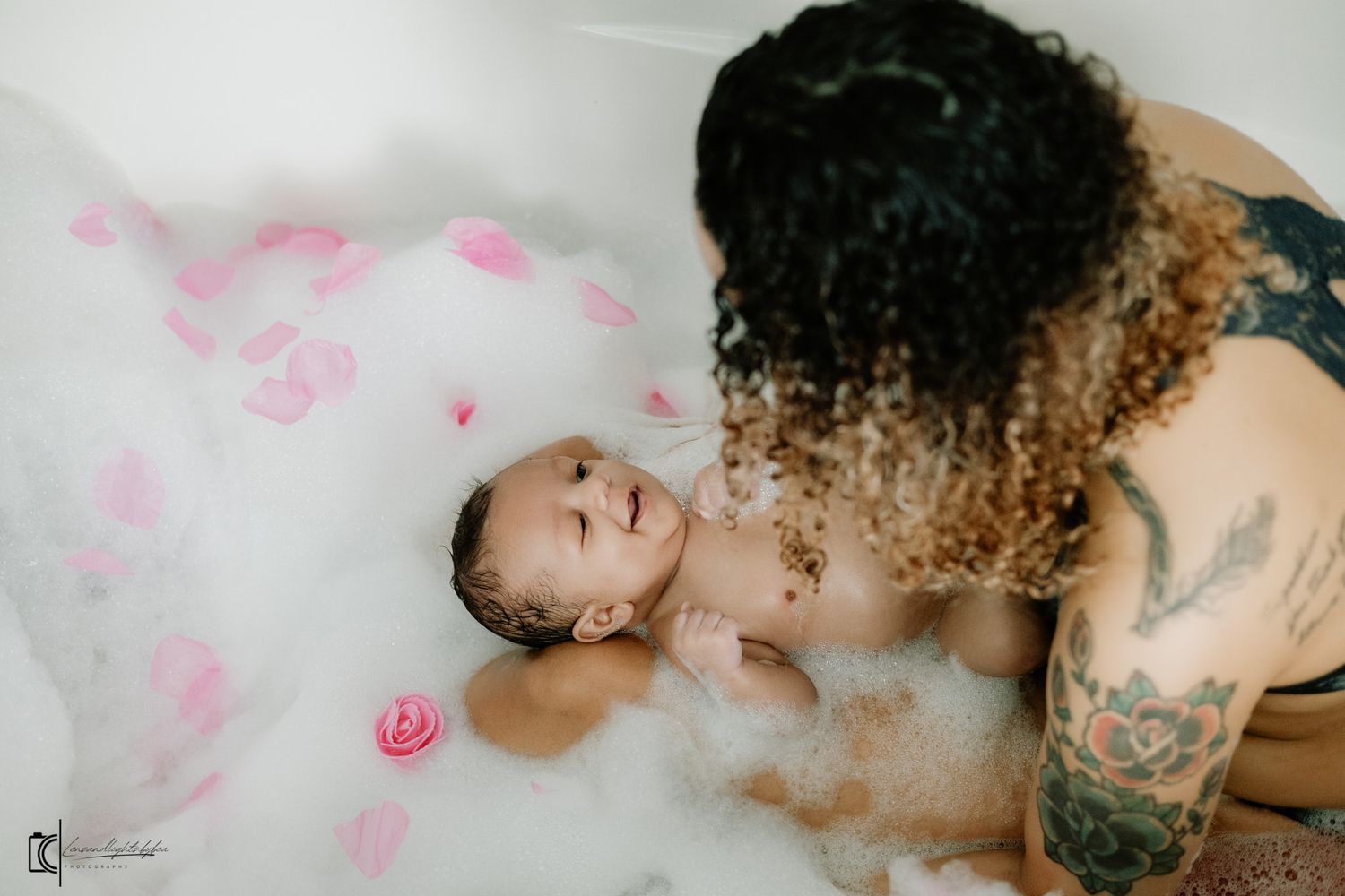 Person with curly hair relaxes in a bathtub filled with pink rose petals and milky water.