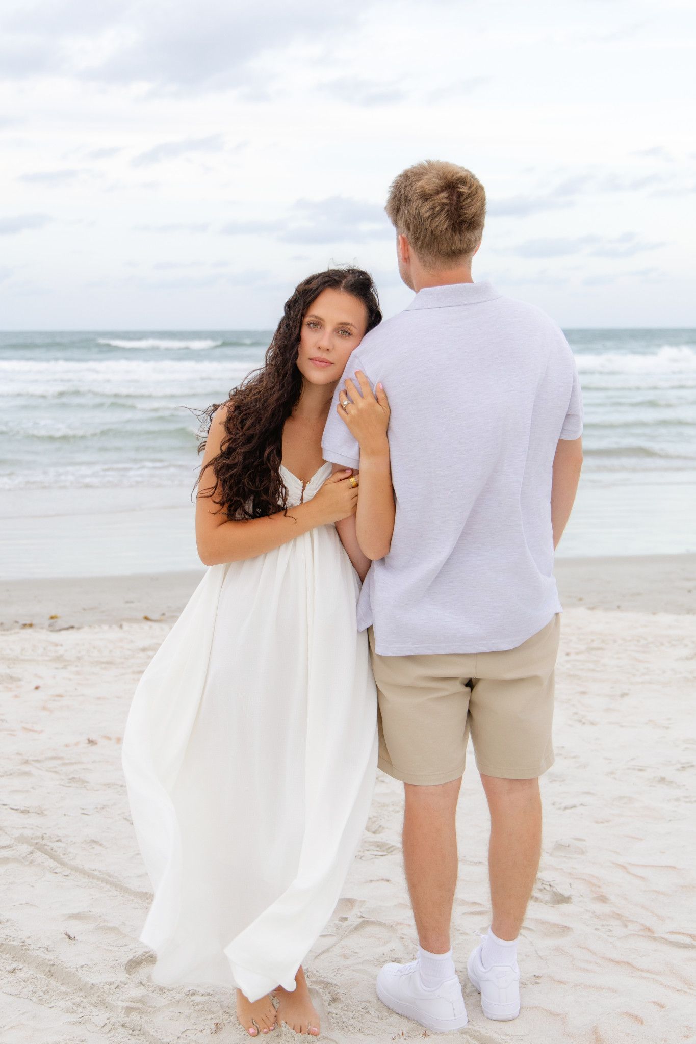 Couple embracing on a sandy beach, woman in white dress leaning against man in casual attire with ocean waves behind.