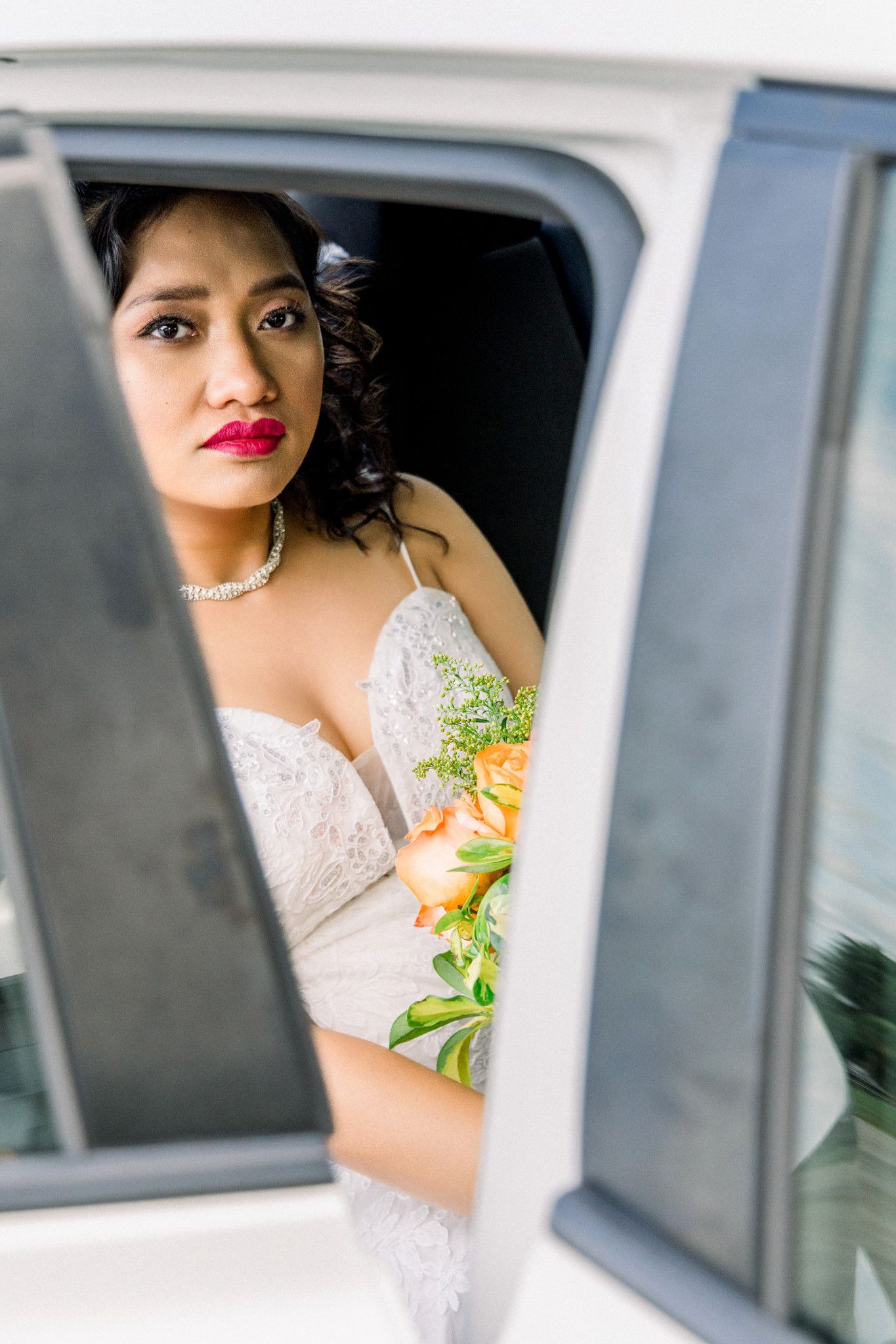Bride in white lace gown with red lips holds peach bouquet, gazing through car window on wedding day.