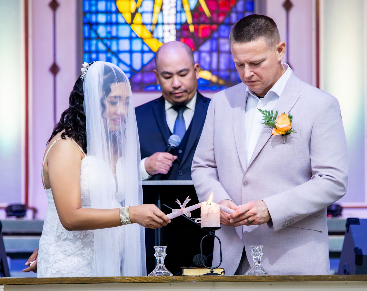 Bride and groom light a unity candle during an indoor church wedding ceremony with stained glass backdrop.