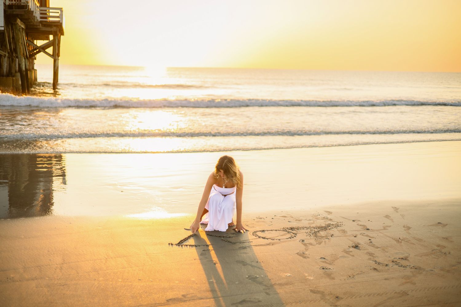 Young girl in white dress drawing in sand at golden sunset on beach.