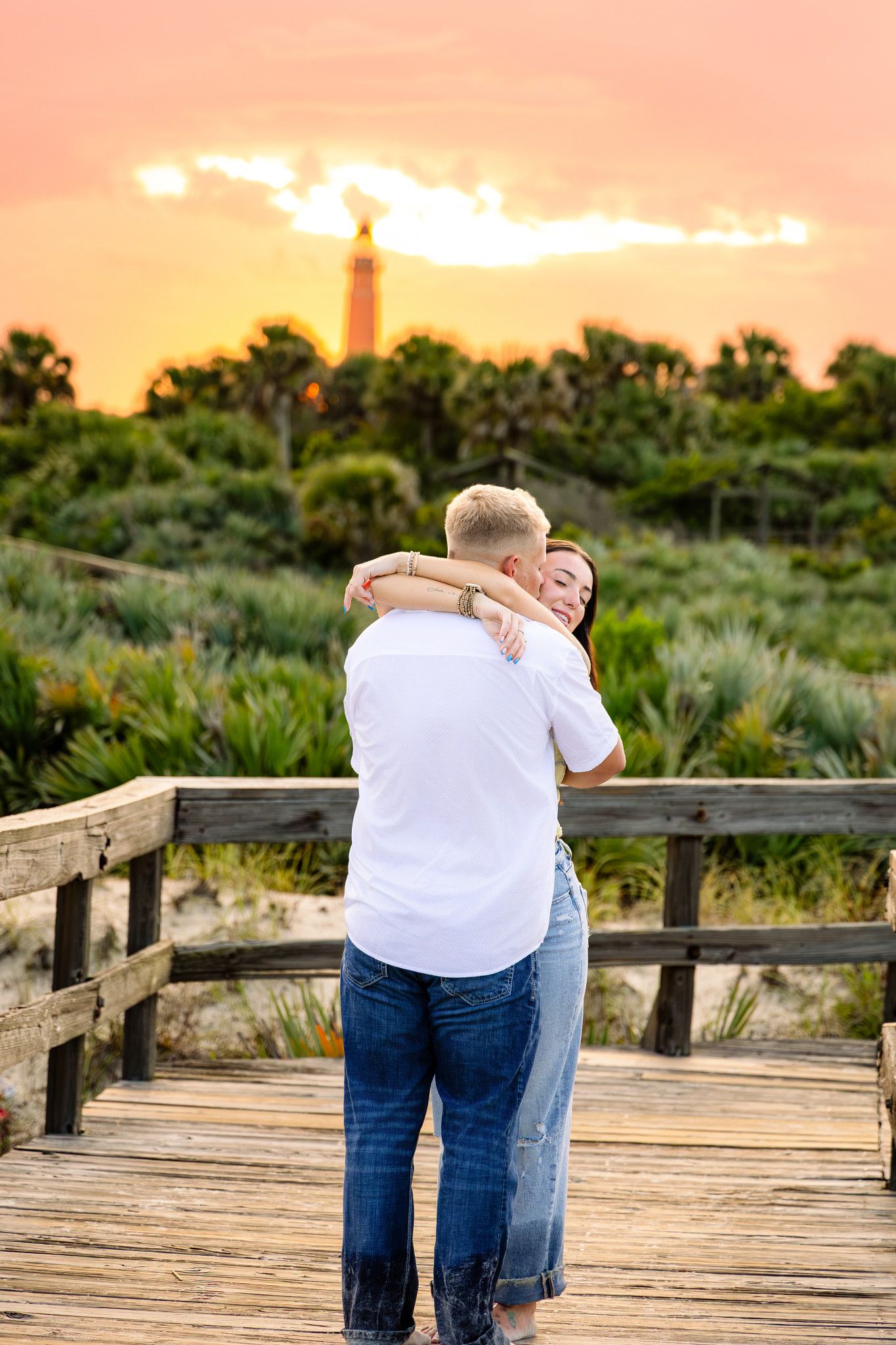 Couple embracing on a wooden boardwalk at sunset with a lighthouse and lush greenery in the background.