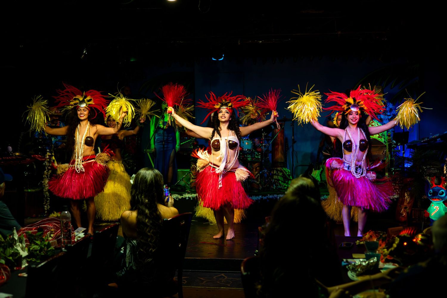 Colorfully costumed performers dance energetically on a dark stage with vibrant lighting effects creating a dramatic atmosphere.