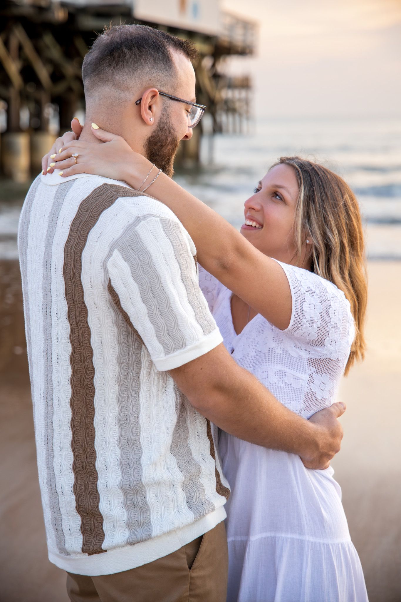 A romantic couple embraces on the beach at sunset near a wooden pier.