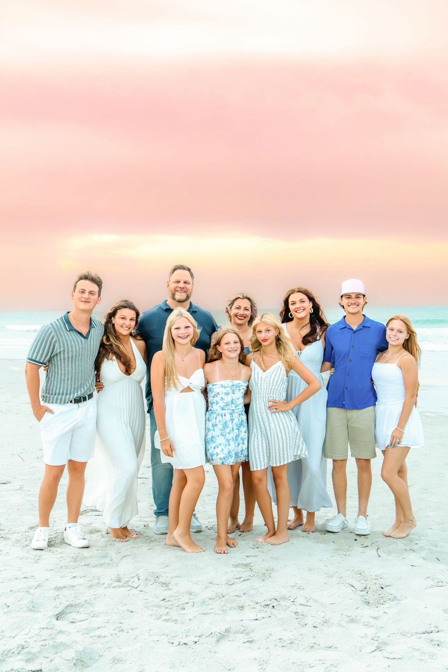 Large family posing together on a sandy beach at sunset with a pink sky, wearing coordinated blue and white outfits.