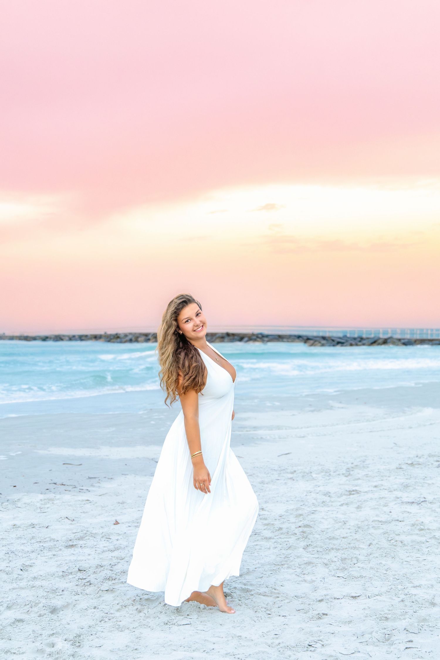Young woman in white maxi dress poses barefoot on sandy beach at pink sunset.