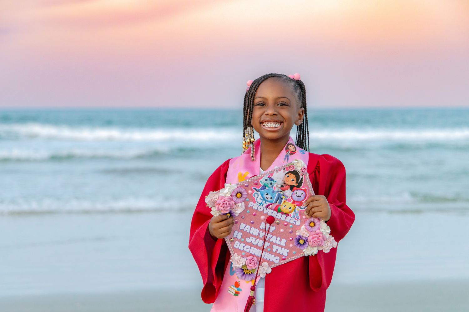 Smiling young girl in red graduation gown holding decorated cap on beach at sunset.
