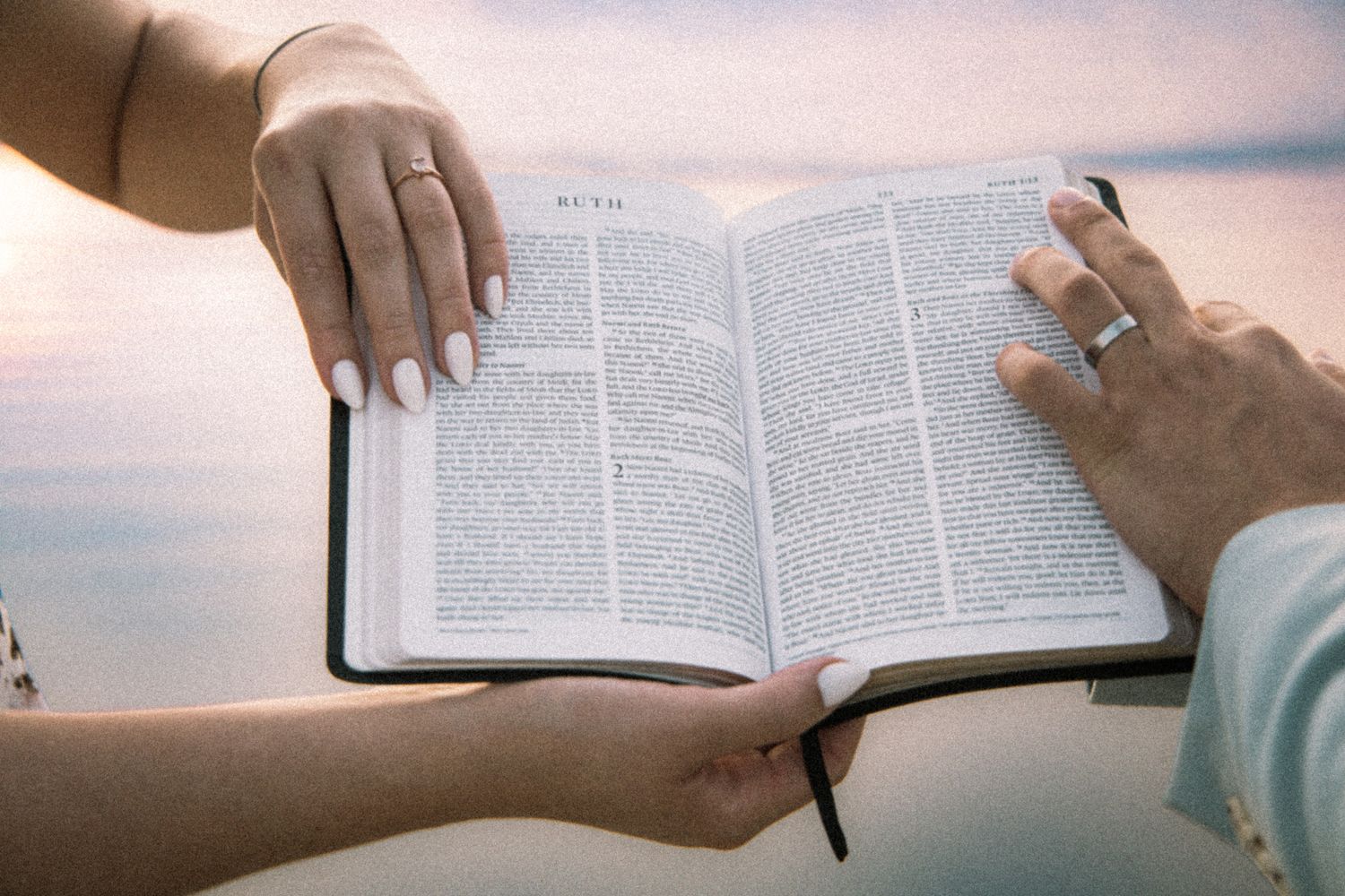 Two people holding an open Bible together outdoors, with a pastel sunset sky in the background.