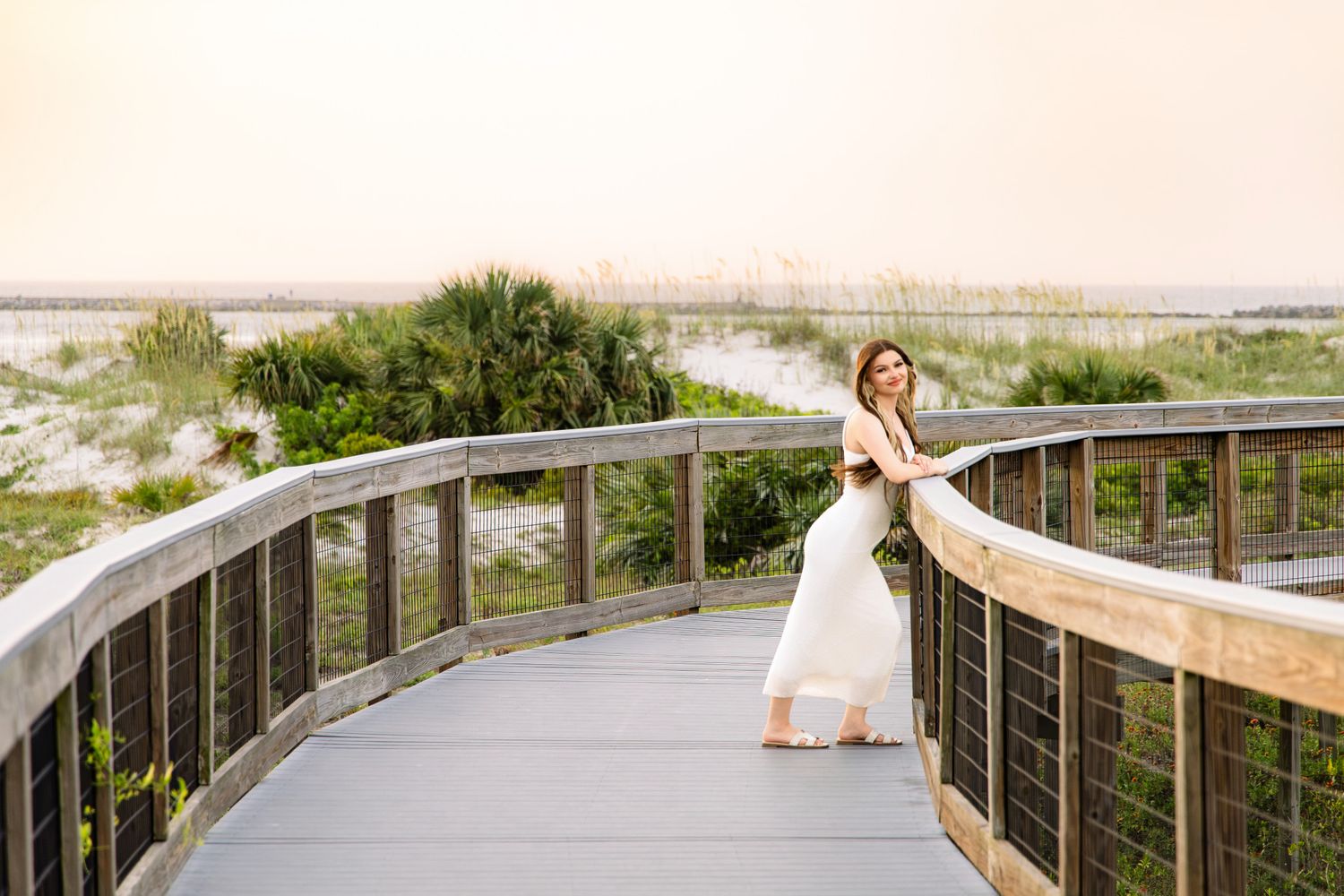 A woman in a white dress poses gracefully on a wooden boardwalk overlooking a sandy beach at sunset.