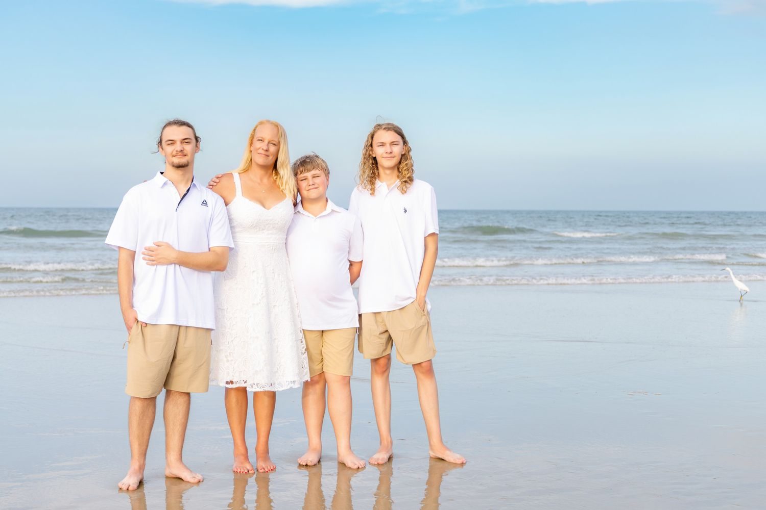 Family of four posing on a sunny beach, wearing white tops and khaki shorts, with ocean waves in the background.
