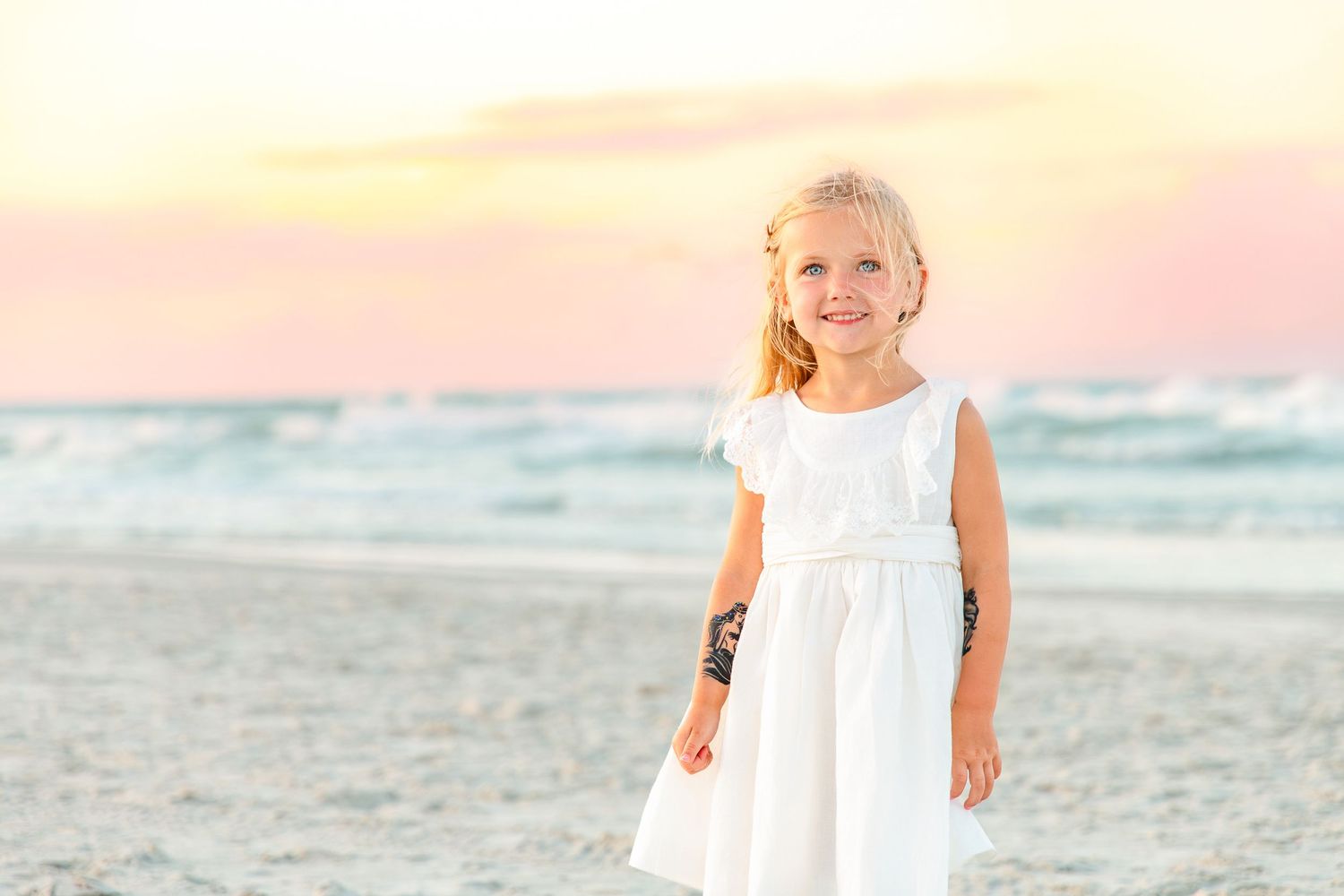 Young girl in white dress smiling on sandy beach at sunset with ocean waves in background.