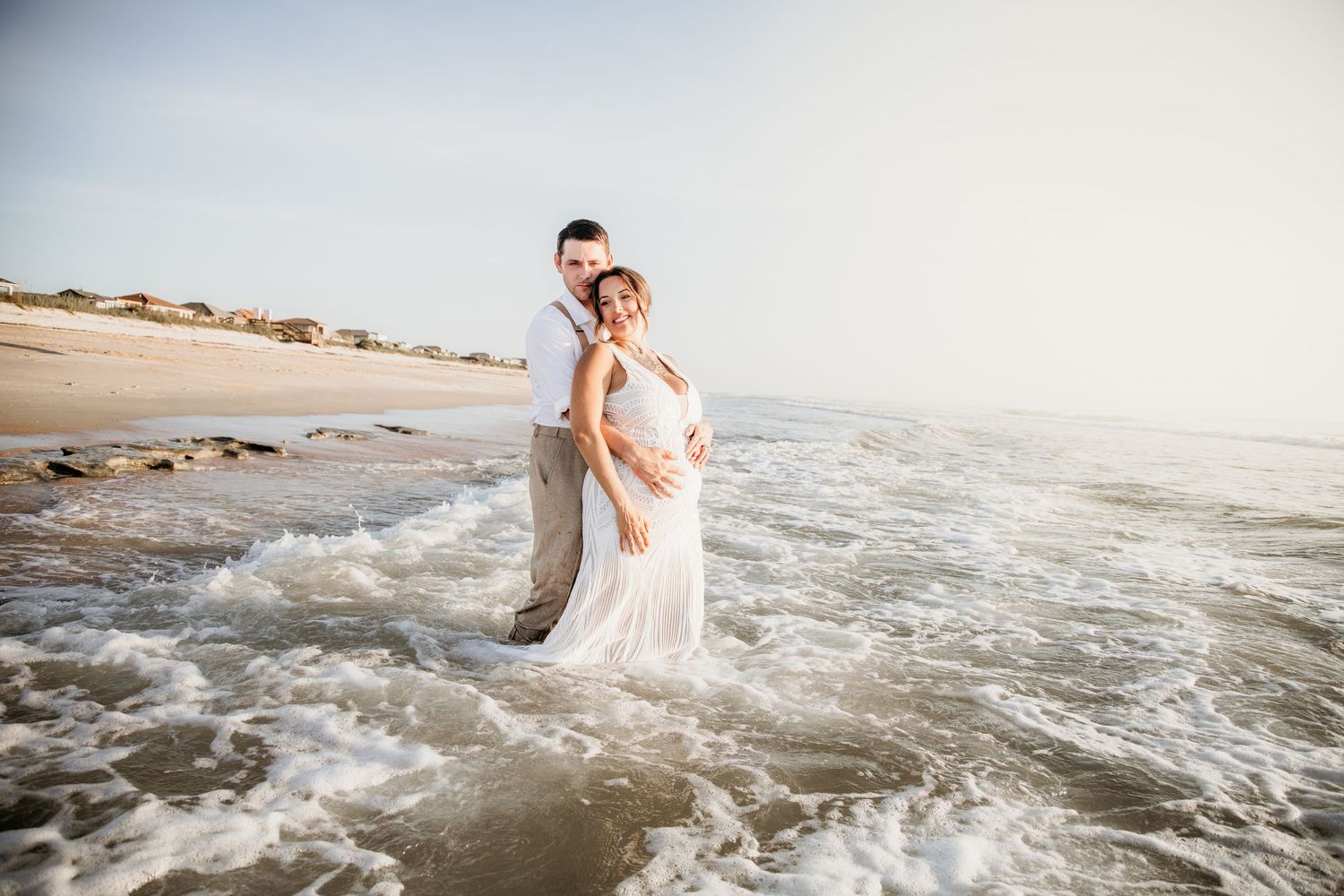 Couple embraces romantically while standing in ocean waves on a sunny beach wedding day.
