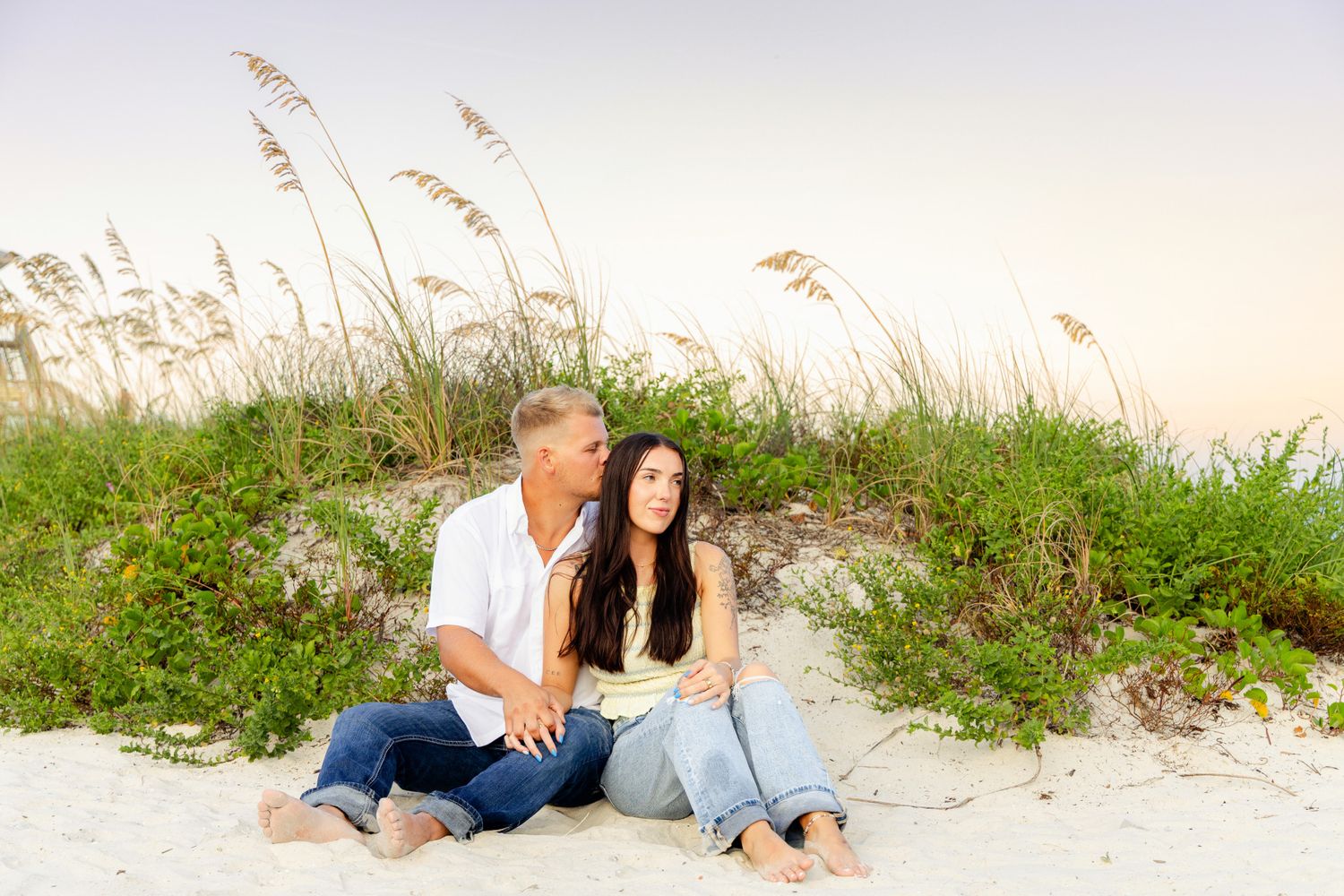 Couple sitting together on white sand dunes surrounded by sea oats during a pastel beach sunset.