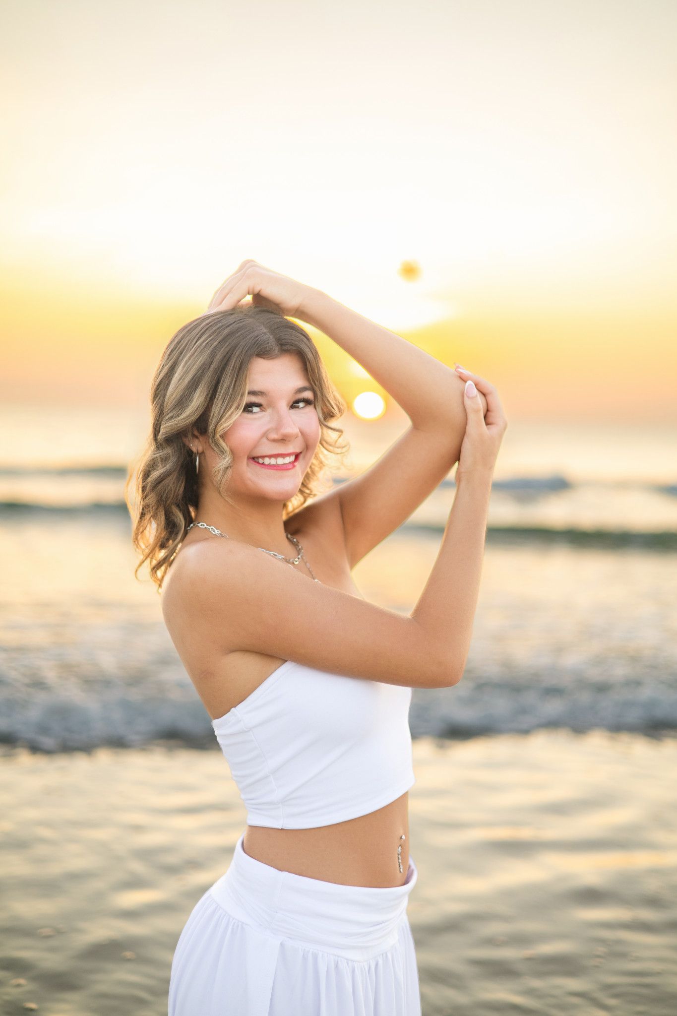 Young woman in white crop top smiling at beach sunset with hands raised above head.