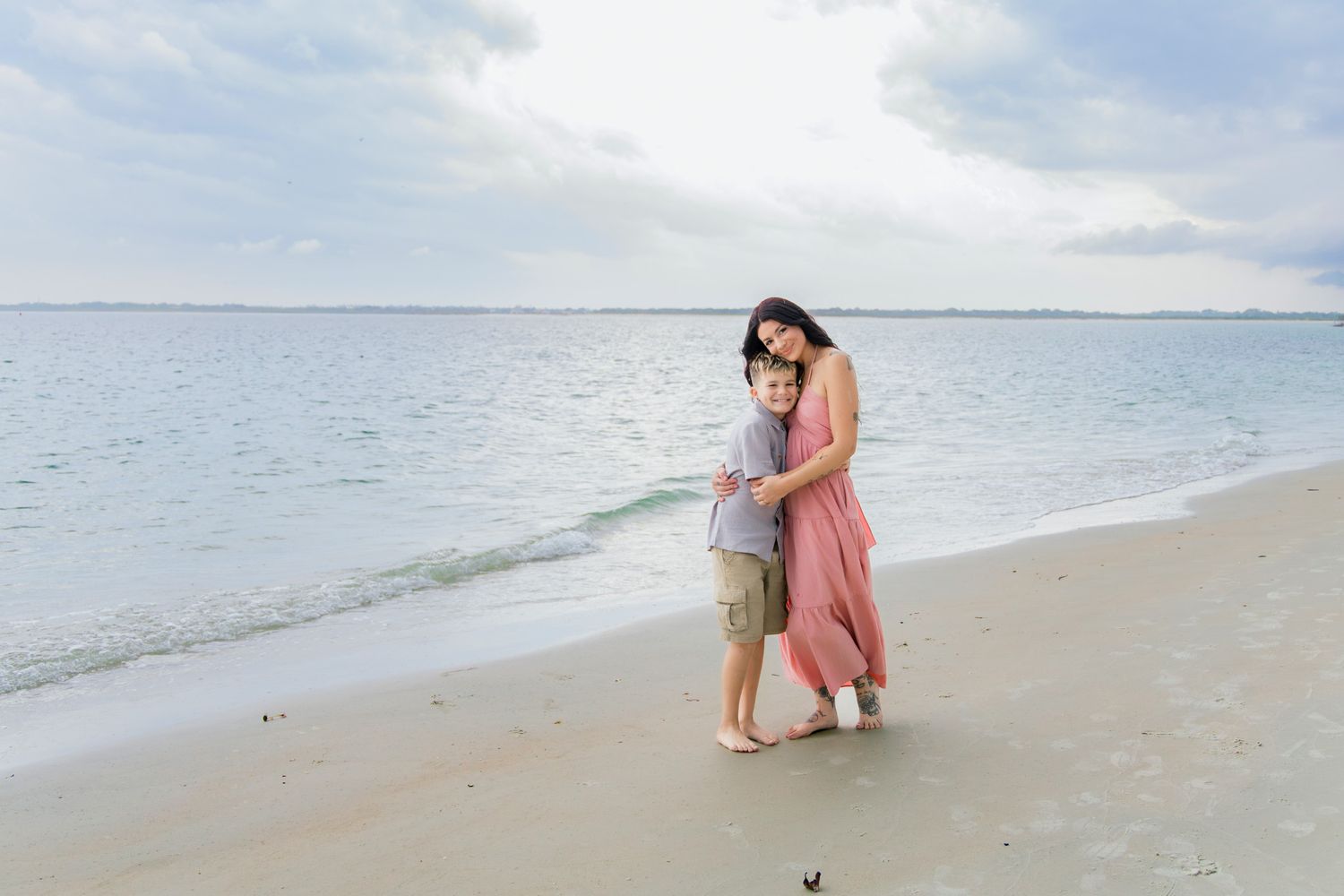Mother in pink dress hugging her young son on a serene beach with gentle ocean waves in the background.