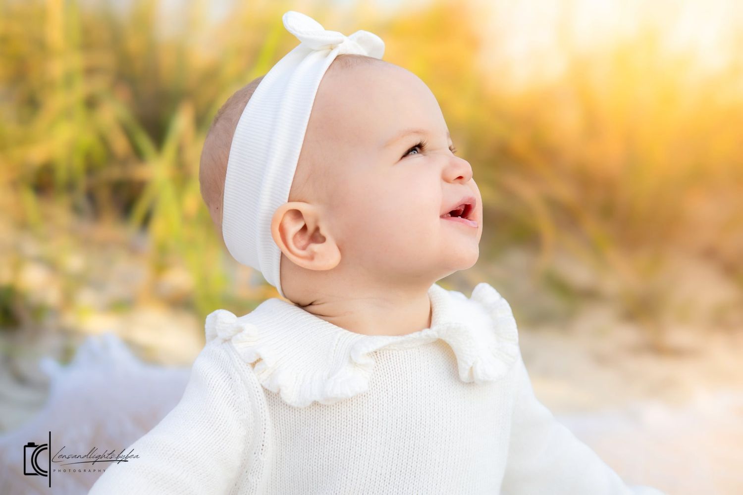 Baby in white outfit sitting in grass during golden hour sunset with warm glowing light in background.