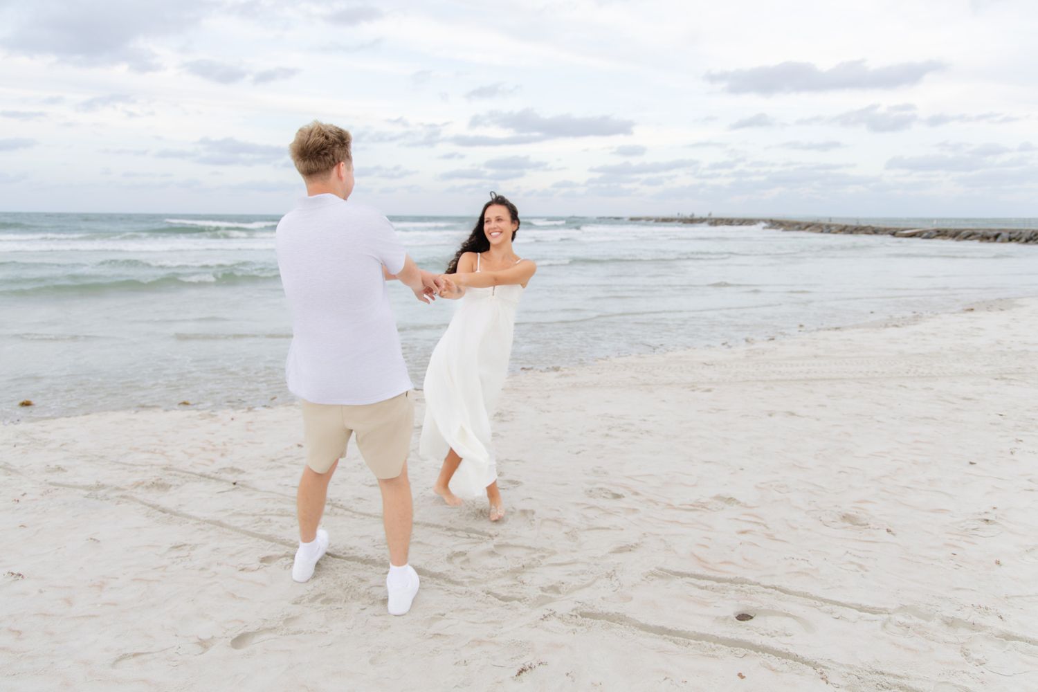 Couple dancing on a sandy beach, woman in white dress, man in casual attire, ocean waves in background.
