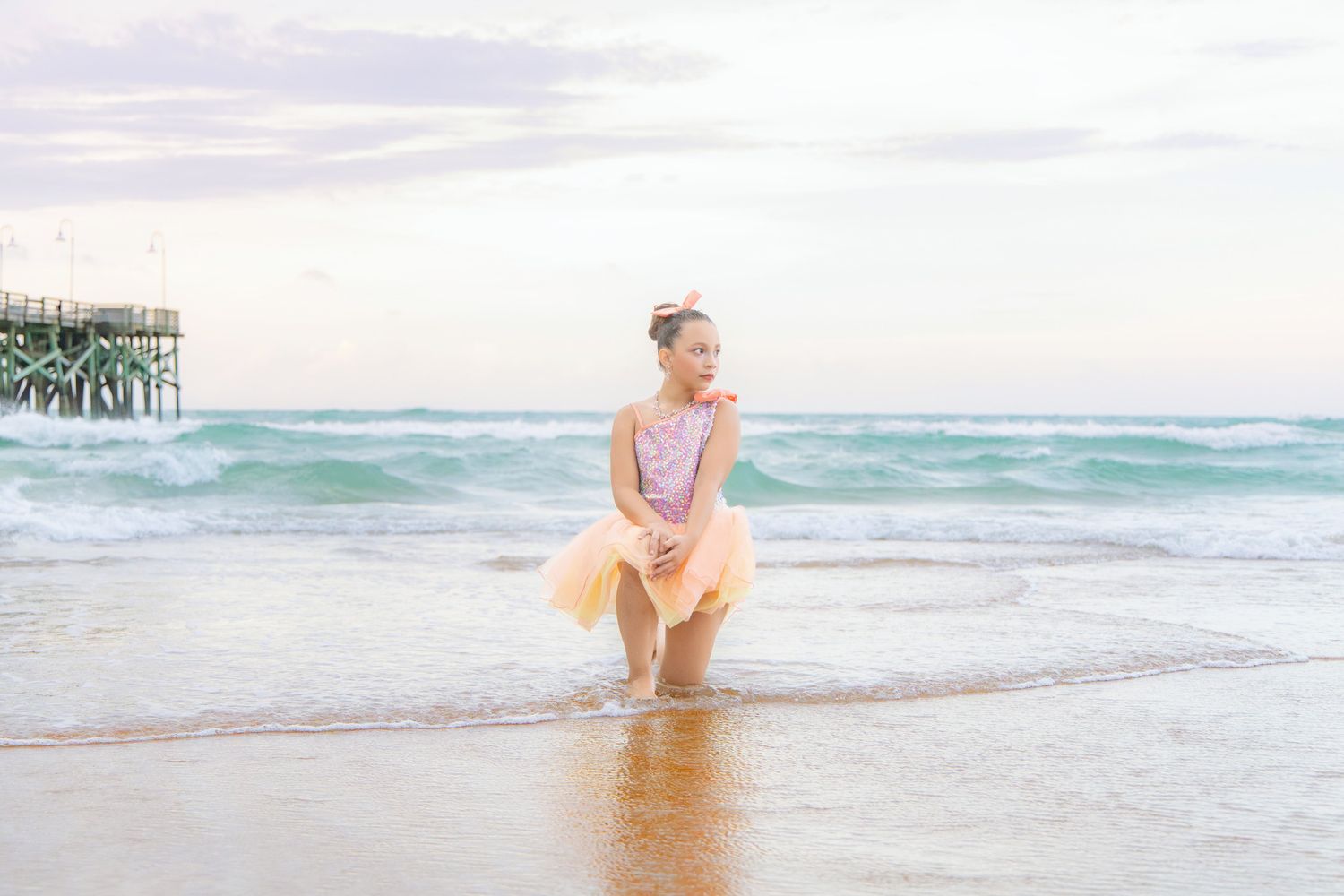 Young girl in pink dress and tutu stands in ocean waves on sandy beach near pier at sunset.