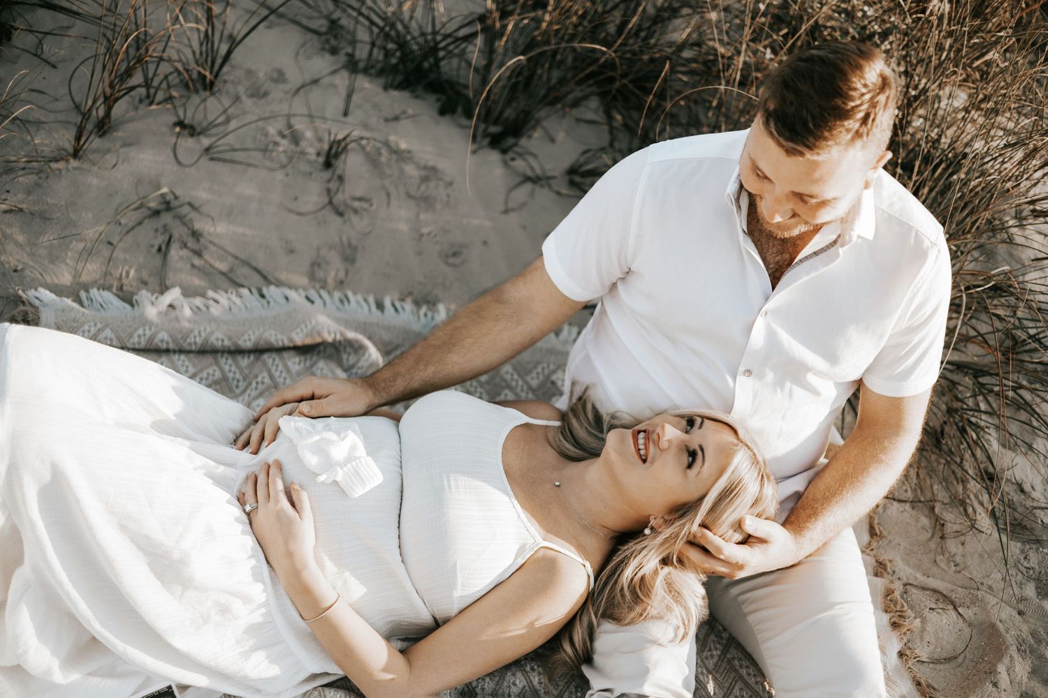 A couple in white outfits poses together on a sandy beach during a romantic photoshoot.