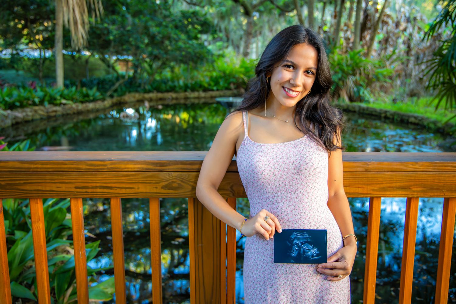 Smiling woman in floral dress holding ultrasound photo on wooden deck surrounded by lush greenery.