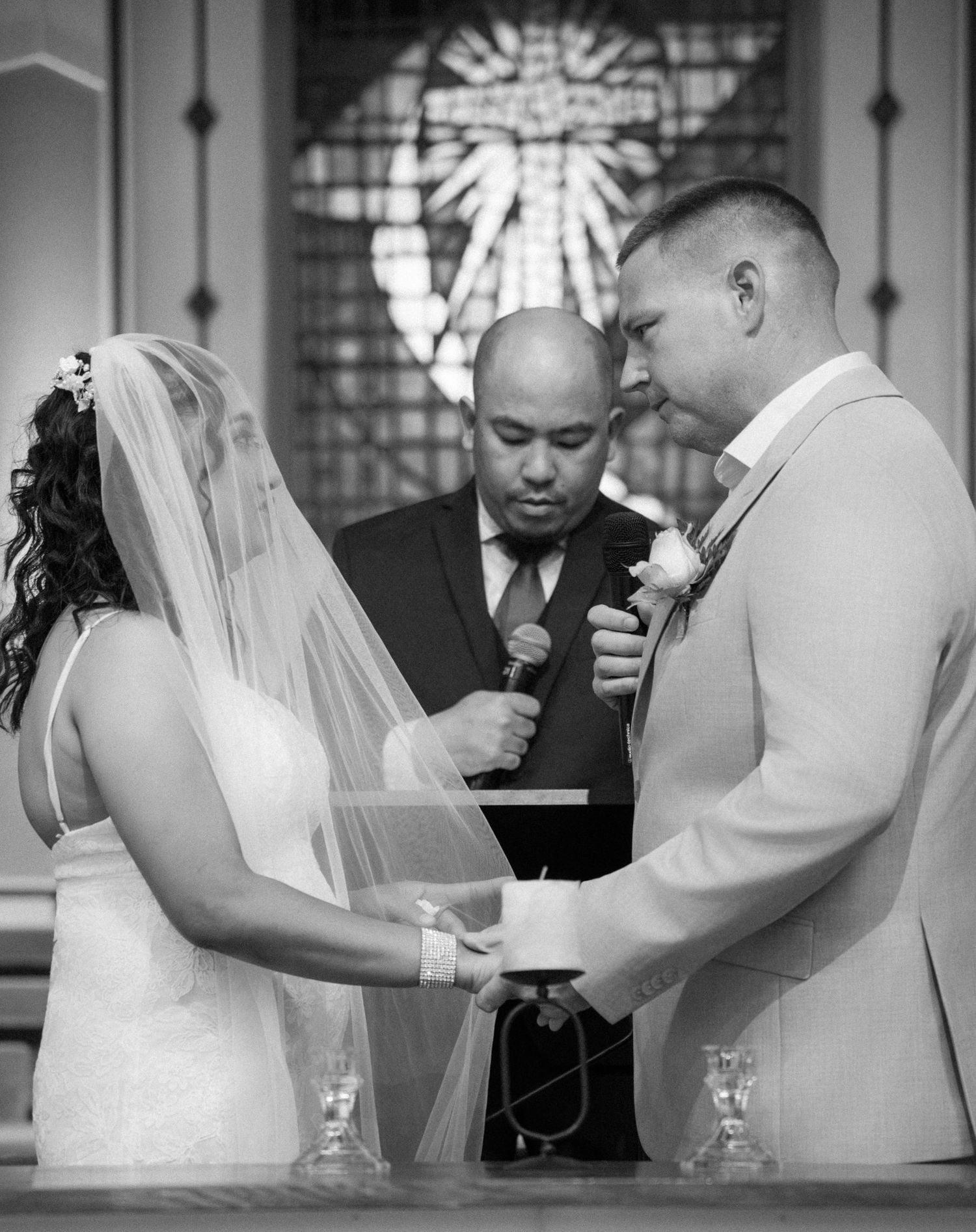 Black and white photo of a bride and groom holding hands during wedding ceremony vows at church altar.