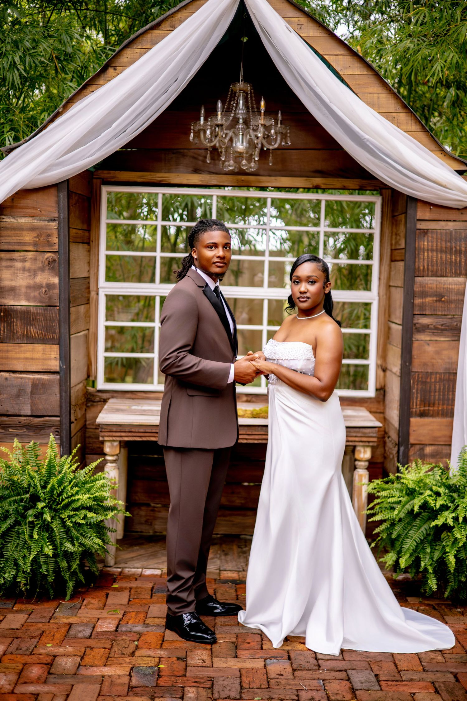 Elegant Black couple in wedding attire pose before rustic wooden chapel with chandelier and white draping.