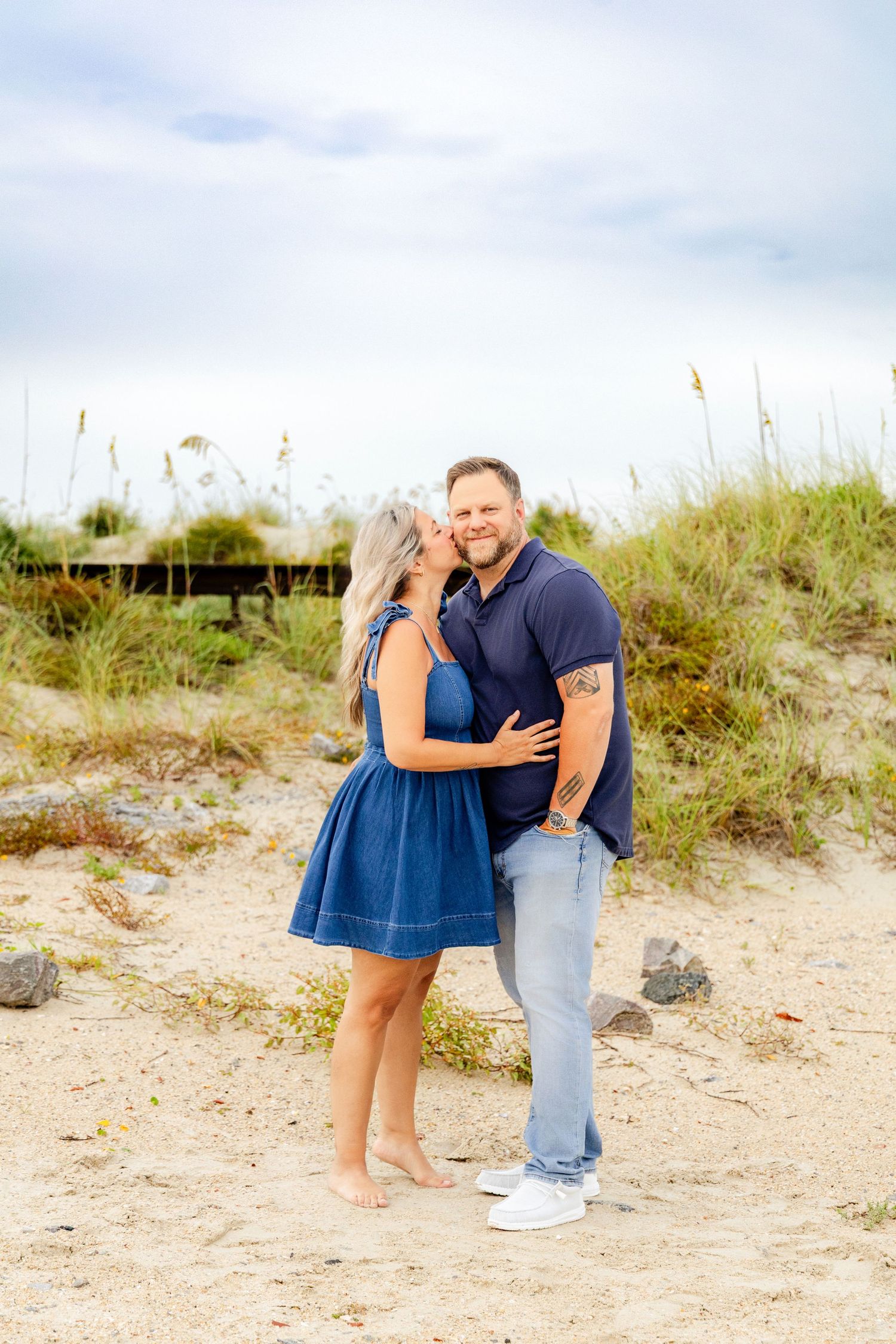 A couple shares a romantic kiss on a sandy beach, woman in blue dress and man in navy polo shirt.