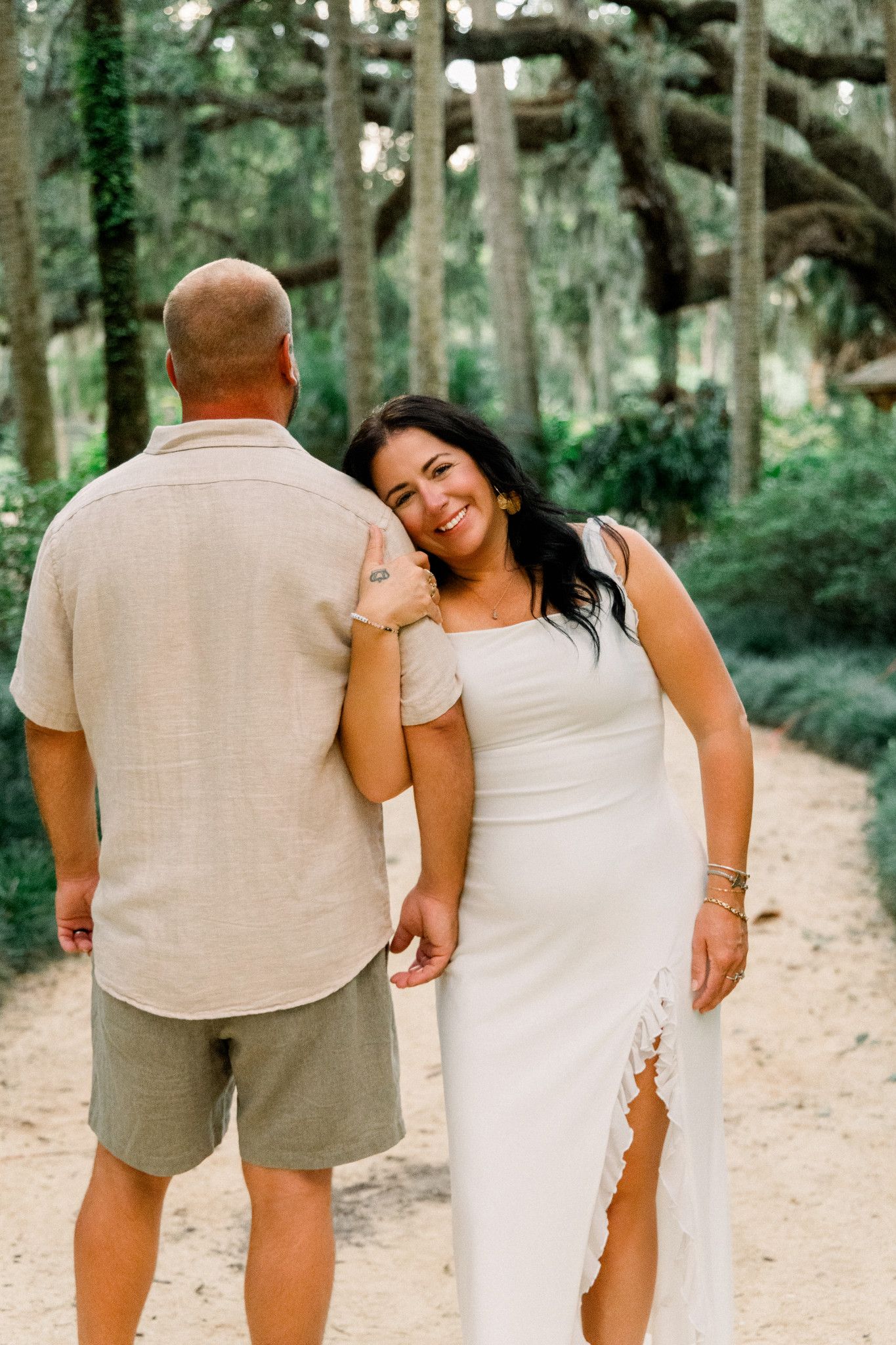 A smiling bride in a white dress leans on her partner's shoulder while holding hands on a sandy forest path.