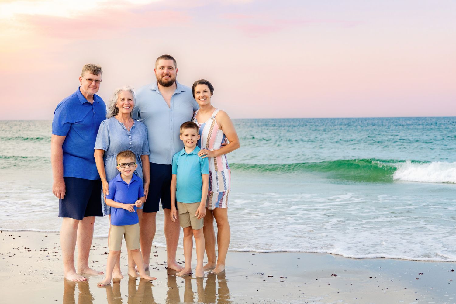 Multi-generational family of six poses together on a sandy beach at sunset with turquoise ocean waves in the background.