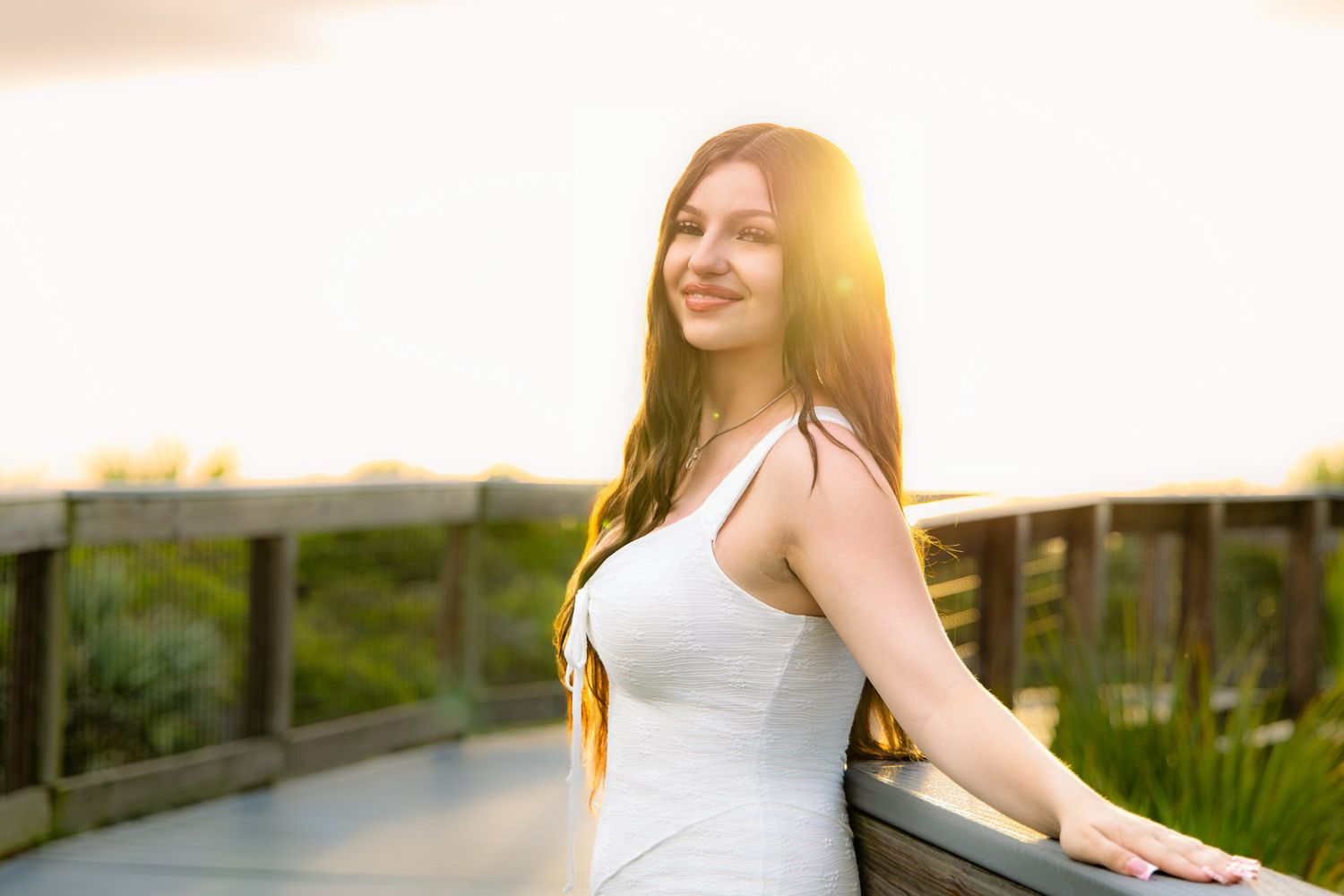 Young woman in white dress leaning on boardwalk railing at golden sunset.
