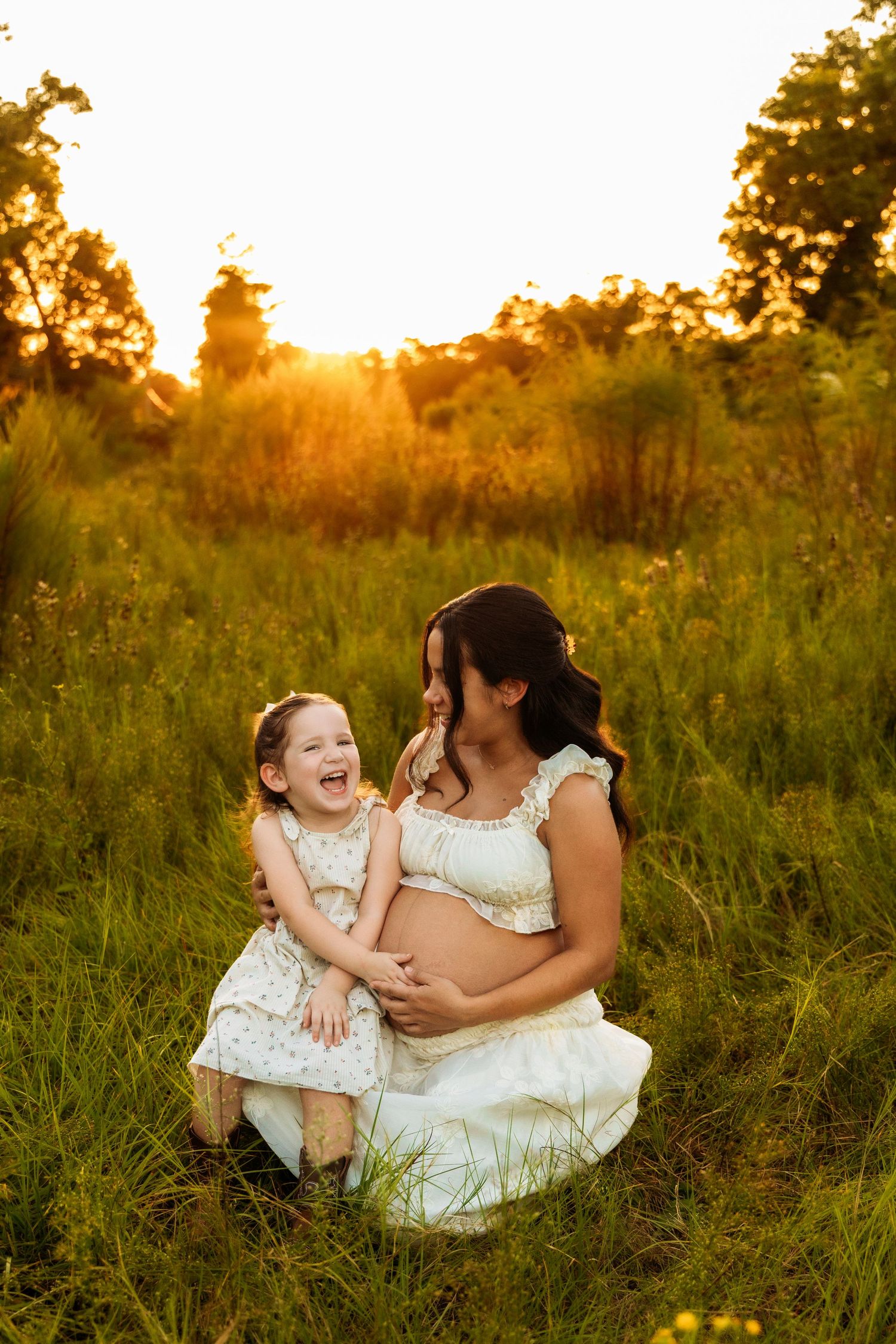 Pregnant mother and laughing toddler daughter sit together in a sunlit green field during golden hour sunset.