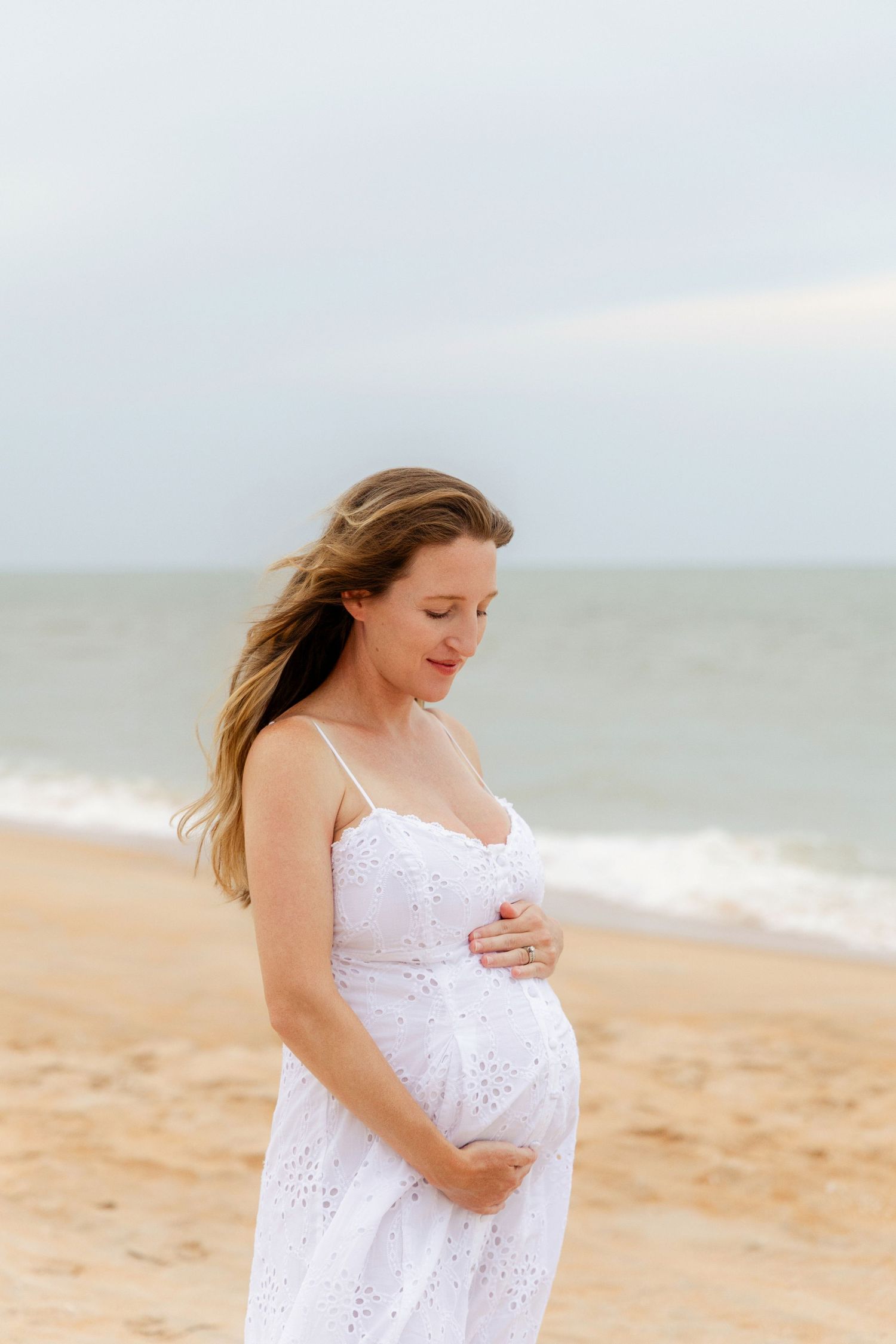 Pregnant woman in white sundress cradling baby bump on sandy beach with ocean waves in background.