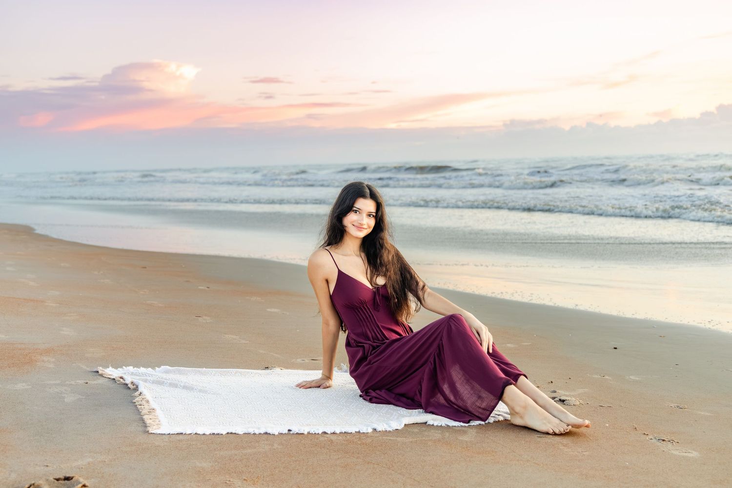 Woman in burgundy dress sitting on white blanket at beach during colorful sunset with ocean waves.