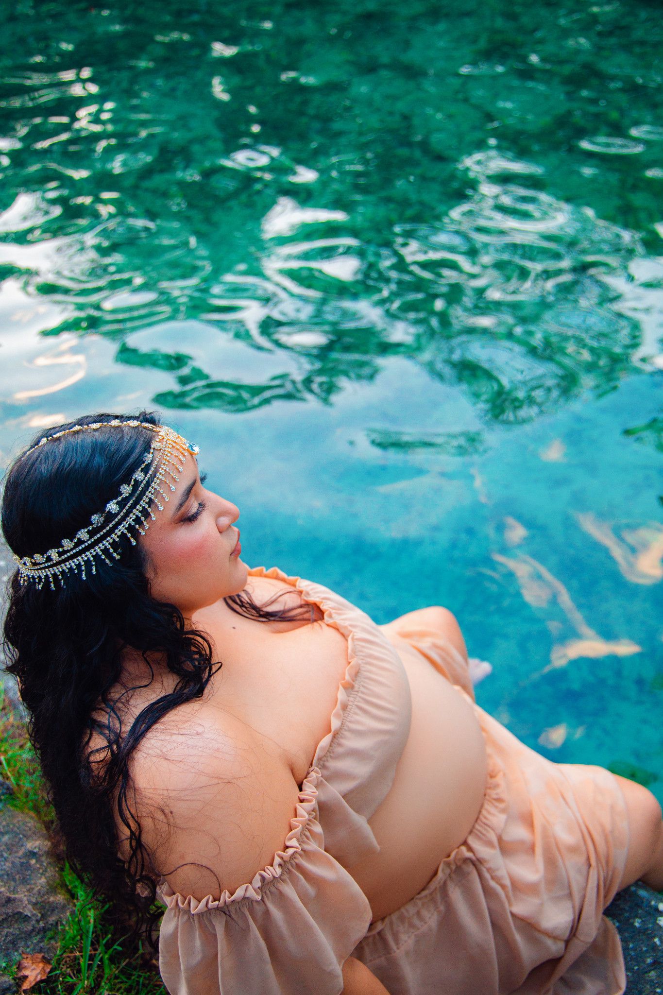 Woman in peach off-shoulder dress with jeweled headpiece sits by turquoise water, eyes closed.